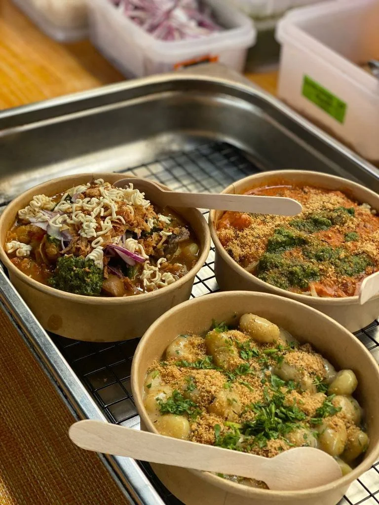 Three bowls of Indian dishes on a wire rack, including gnocchi with sauce and herbs, vegetable curry with flattened rice topping, and a lentil or chickpea dish garnished with cilantro, with wooden spoons for serving.