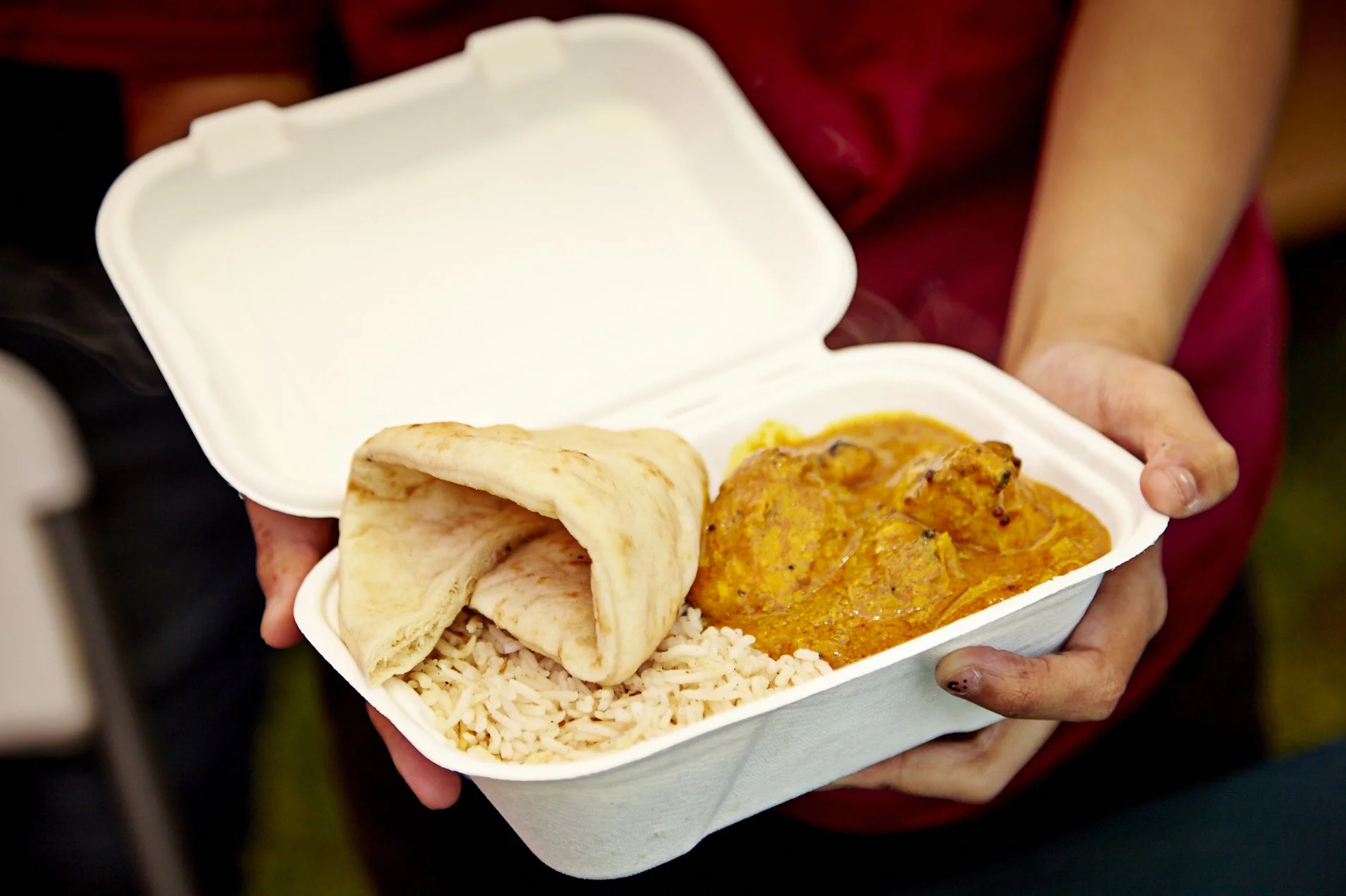 A person holding a white food container with rice, naan bread, and chicken curry.