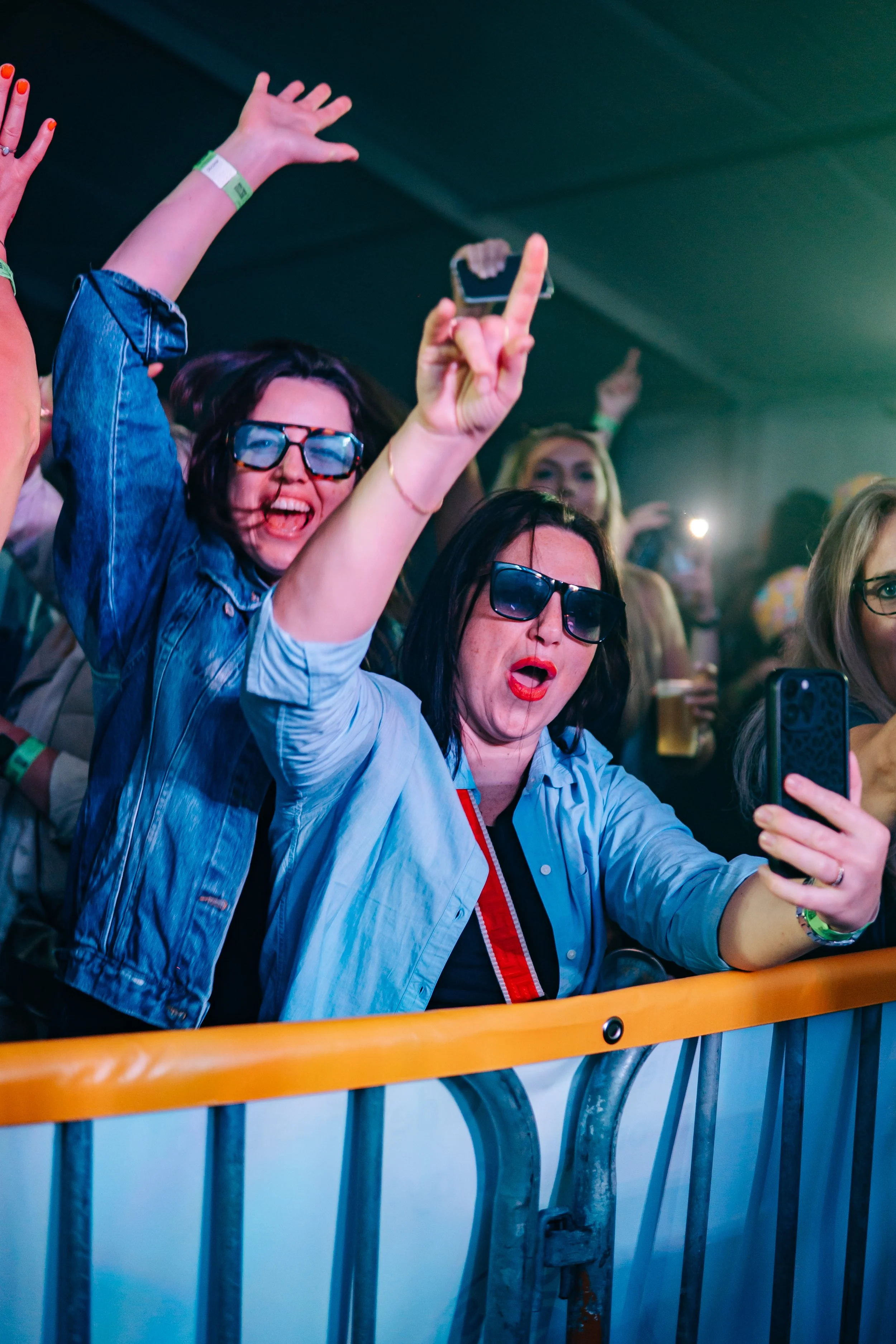 Two women at a concert or event, wearing sunglasses, enthusiastically taking selfies and dancing behind a barrier with orange and gray metal railing, with other attendees in the background.