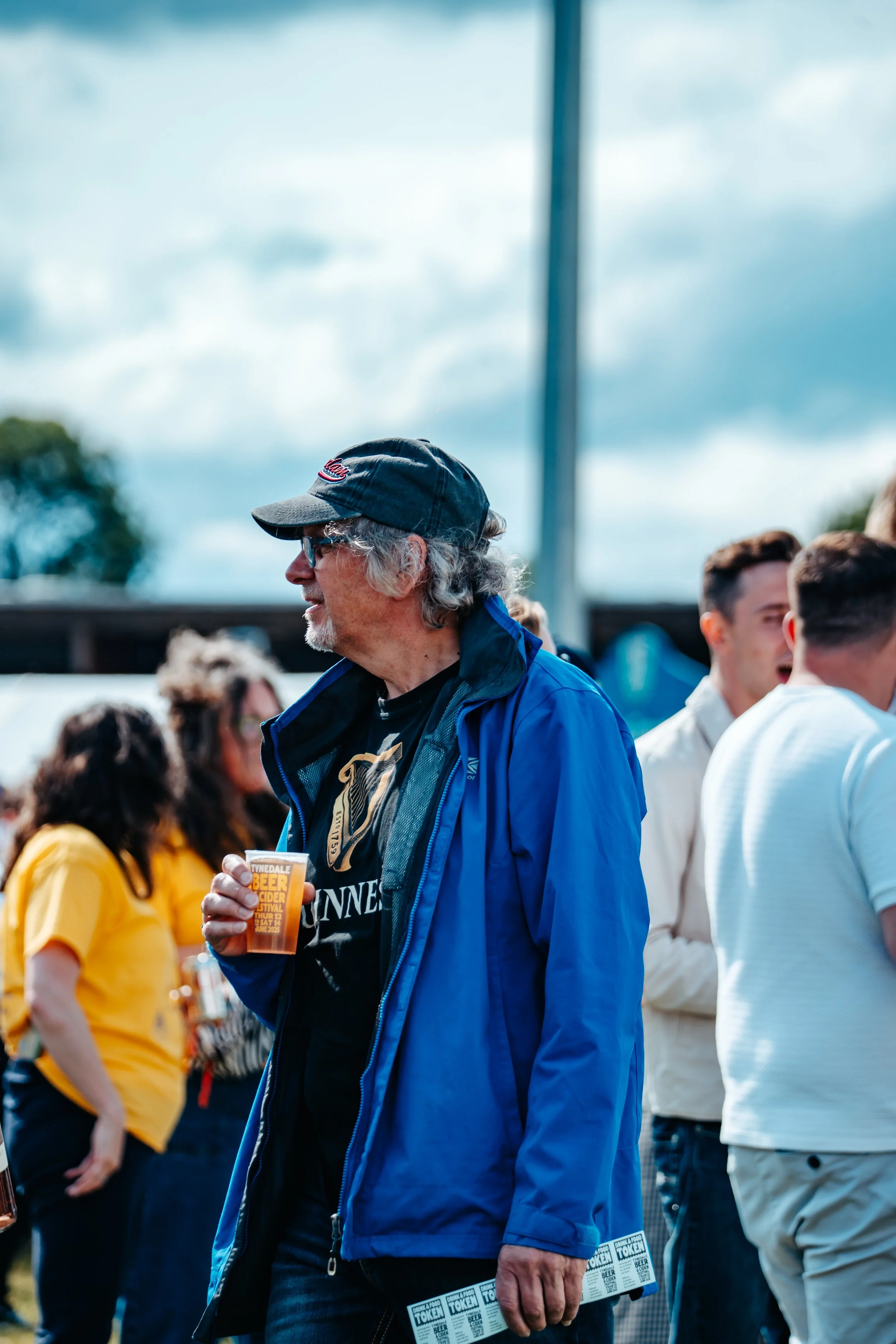 A man with gray hair, glasses, and a black cap holding a drink at an outdoor event with people in the background.