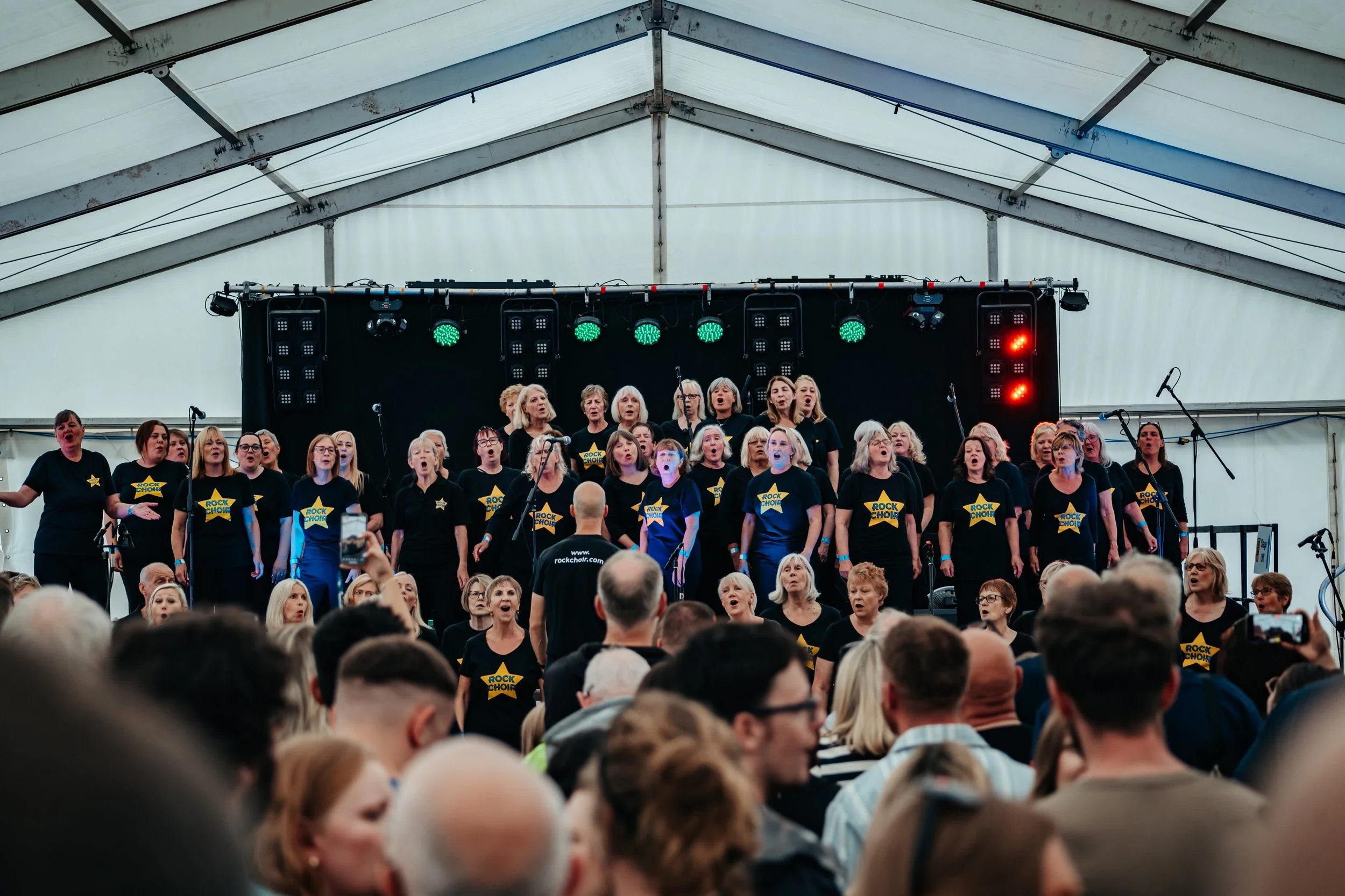 A choir performance under a large white tent, with many singers wearing black shirts labeled 'ROCK CHOIR' and a crowd watching.