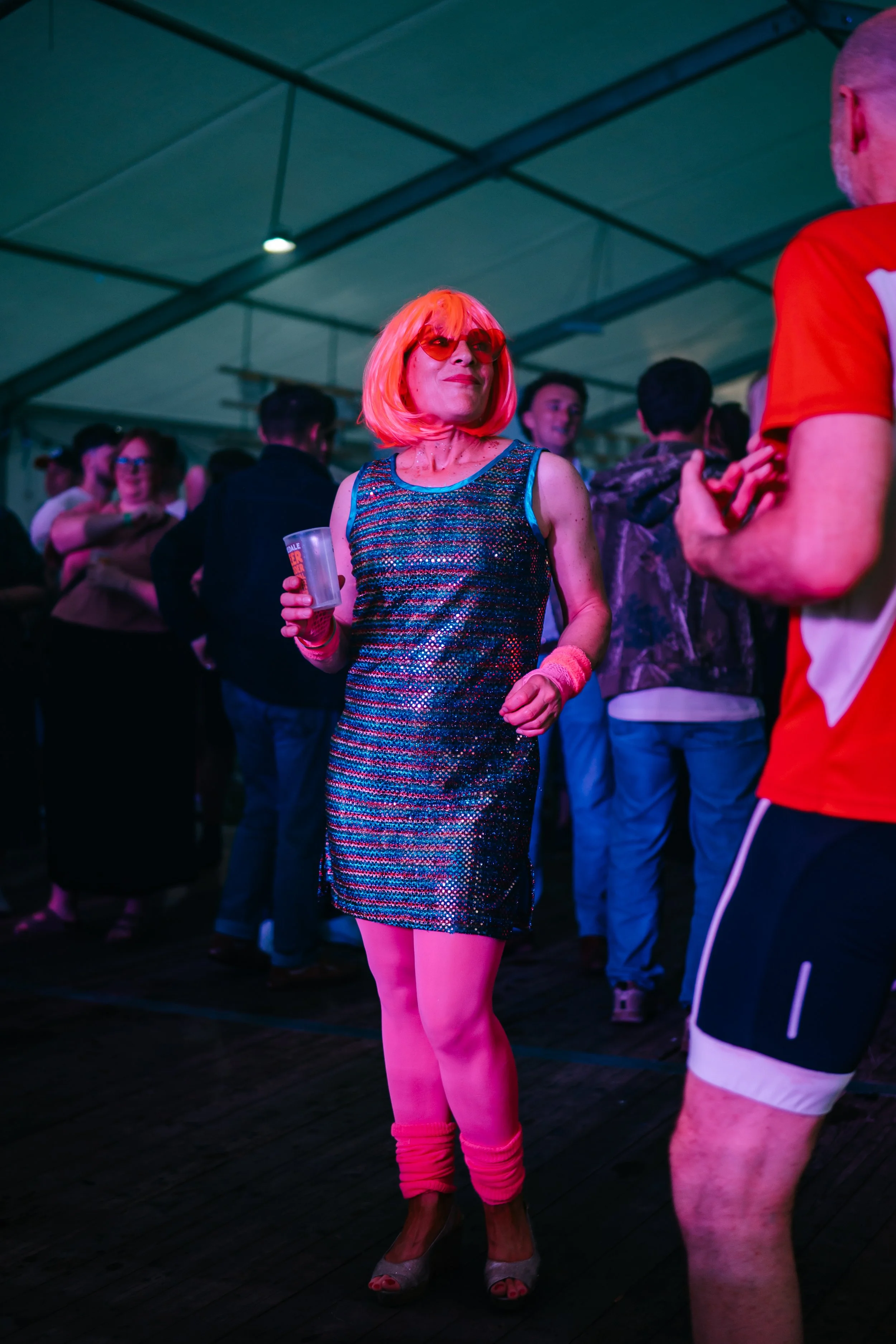 A woman with orange hair, sunglasses, and a shiny dress dancing in a crowded indoor party or dance event.