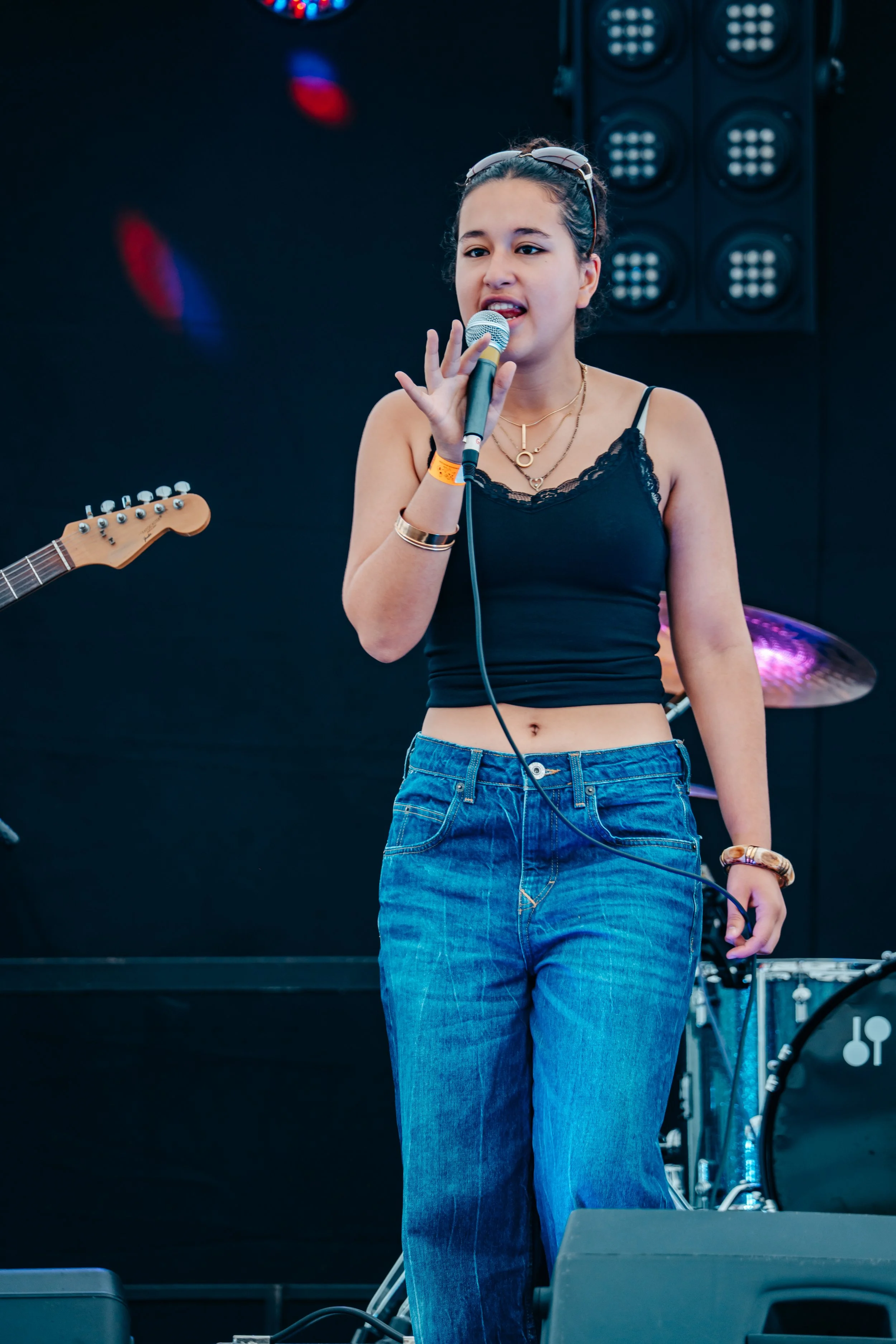 Young woman singing into a microphone on stage, wearing a black spaghetti strap top, blue jeans, with musical equipment and stage lights in the background.