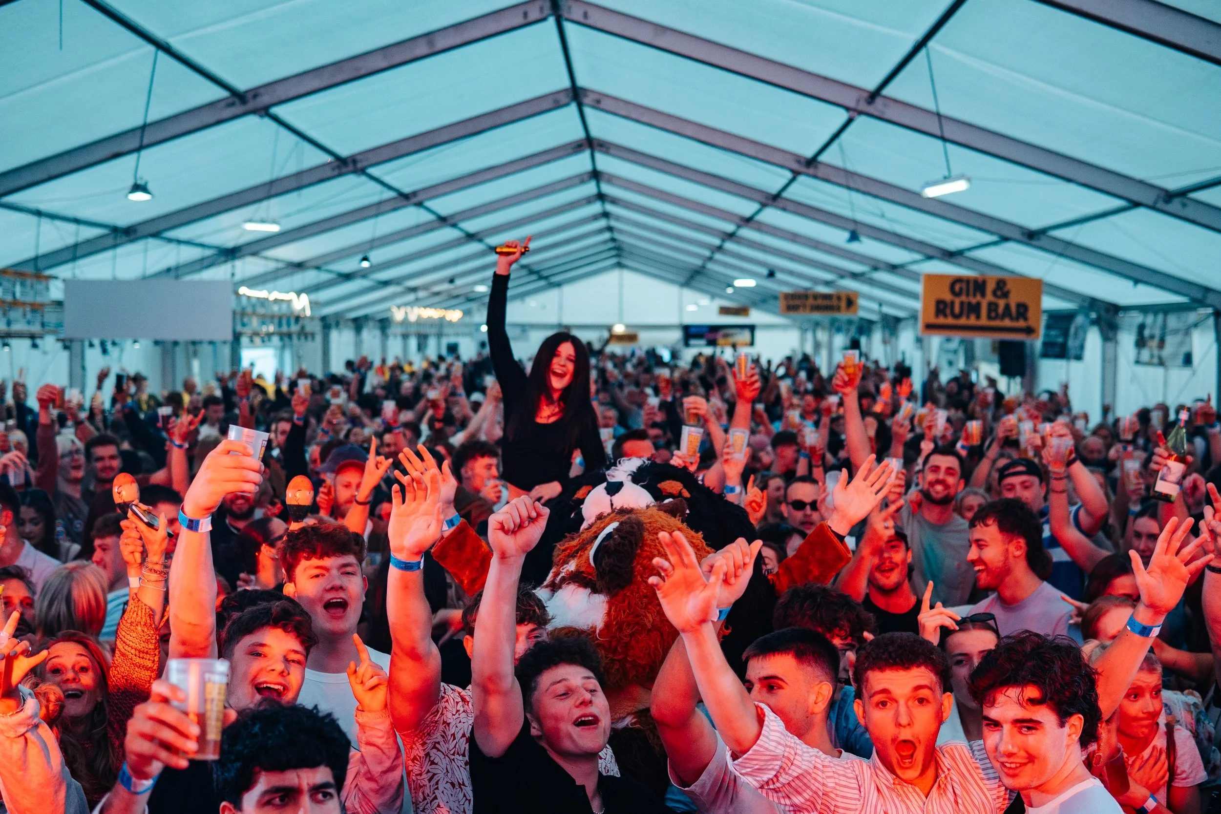 Crowd of people inside a large tent at a festival or party, cheering and holding drinks, with a woman sitting on someone's shoulders holding a drink up and a person in a lion costume in the crowd.