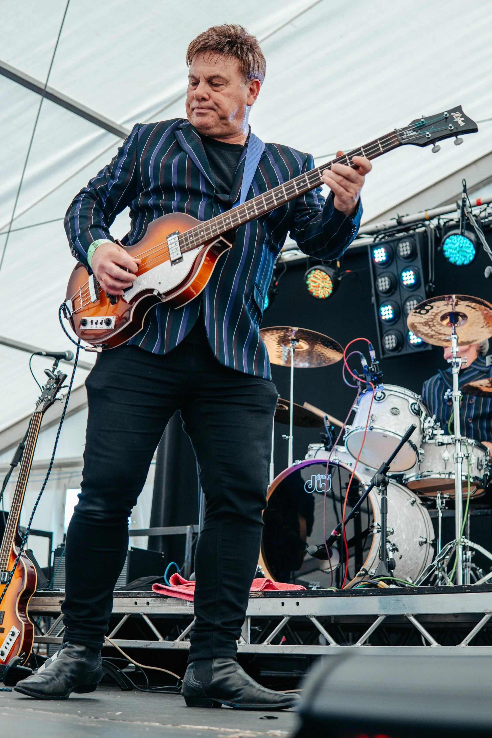 A male musician playing an electric guitar on stage during a concert, with drums and stage lighting in the background.