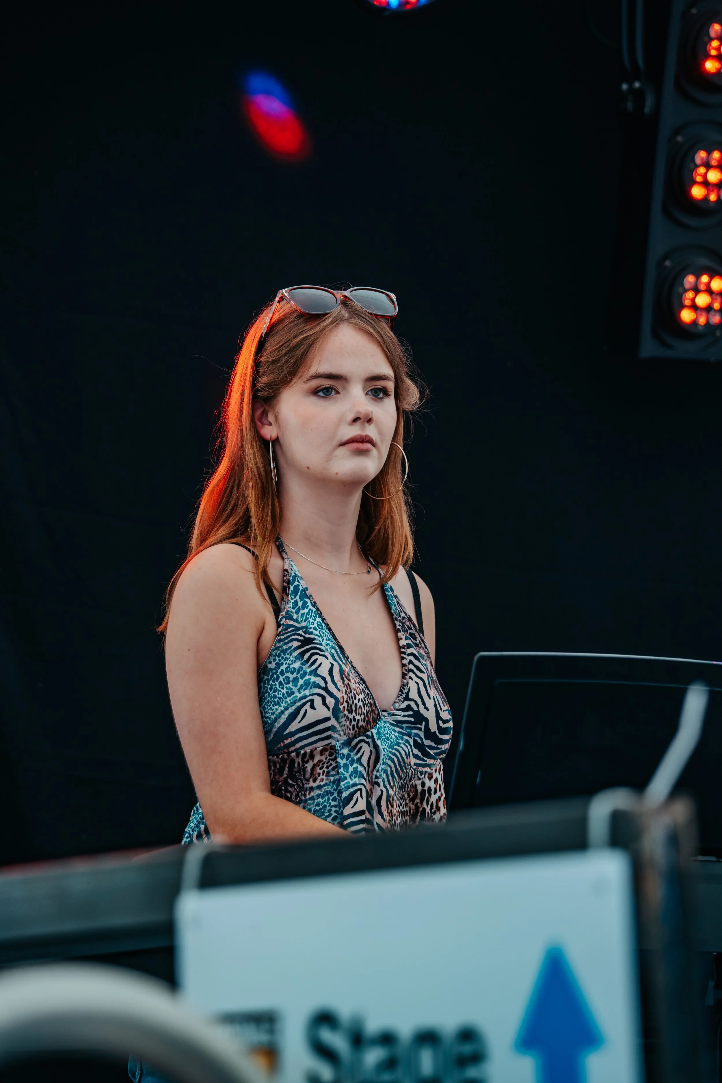 A young woman with red hair, wearing a sleeveless animal print top and sunglasses on her head, standing in front of a black background with stage lights to the side.