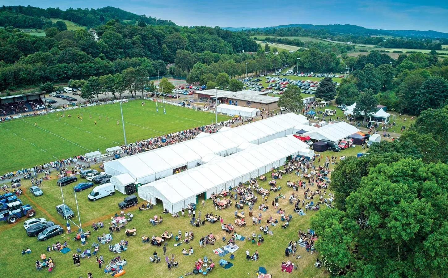 Aerial view of a sporting event at a park with a rugby or football field, surrounded by white tents and crowds of people sitting and standing on the grass, with a line of parked cars and trees nearby.