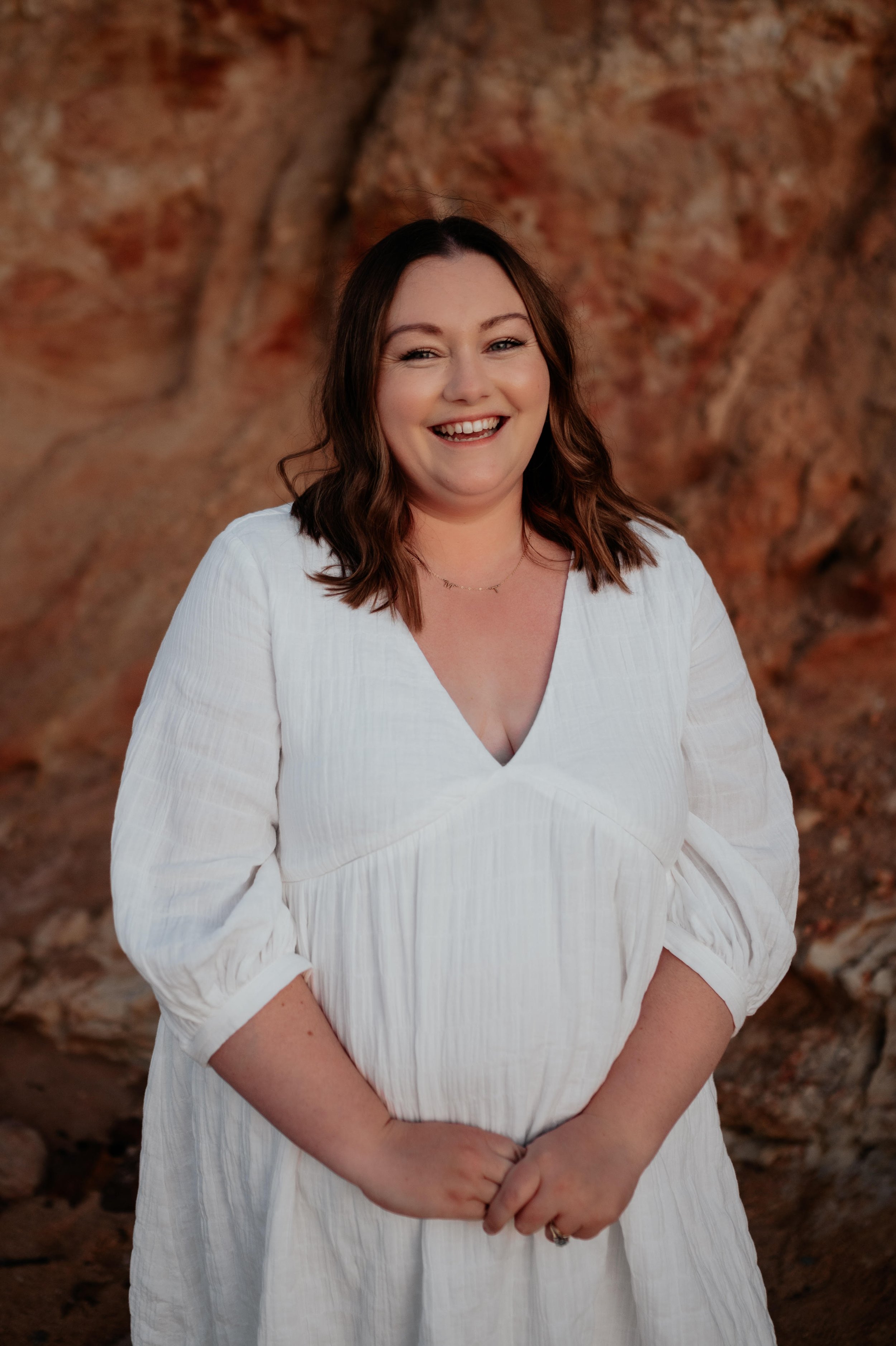 A woman with shoulder-length brown hair smiling, wearing a white dress with rolled-up sleeves, standing outdoors in front of a rocky background.