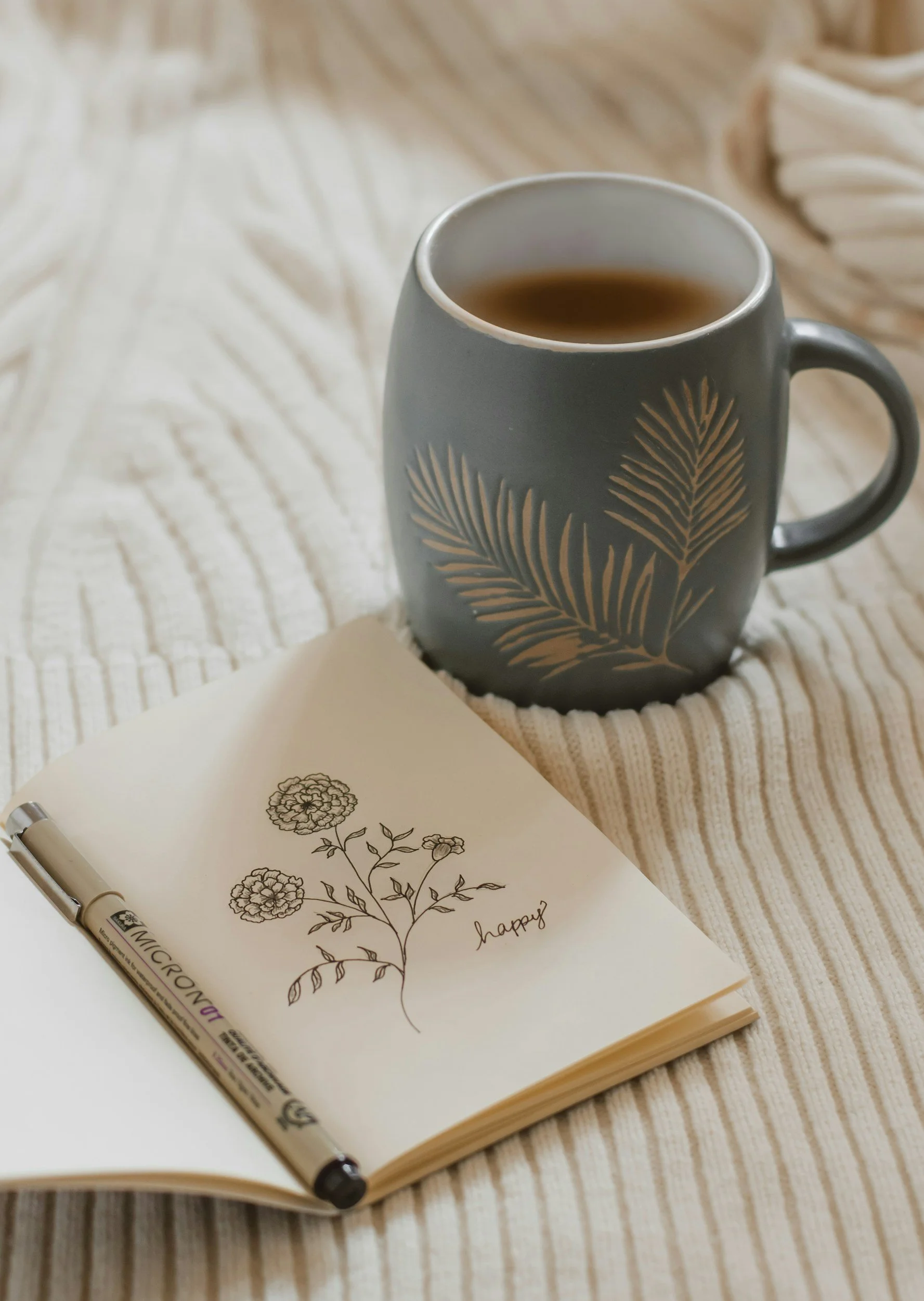 A cup of tea on a beige, textured blanket with a sketchbook and pen nearby, the sketchbook has a drawing of flowers and the word 'happy' written on it.