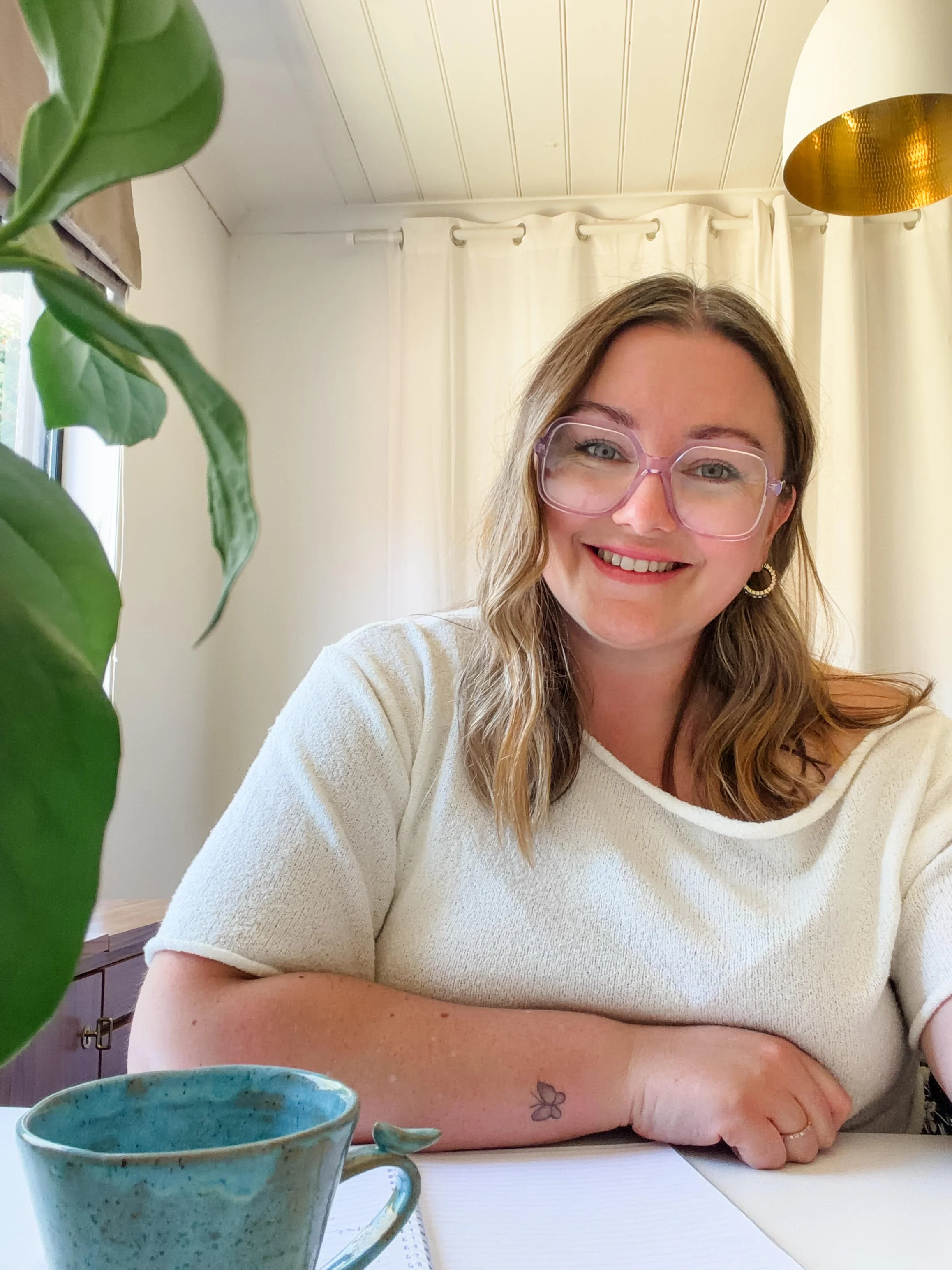 A woman with shoulder-length wavy hair, wearing pink glasses, gold hoop earrings, and a cream-colored top, smiling while sitting at a table with a turquoise mug, a notebook, and a pen. She has a butterfly tattoo on her left forearm. The background features cream curtains and a white ceiling with a gold-colored light fixture.