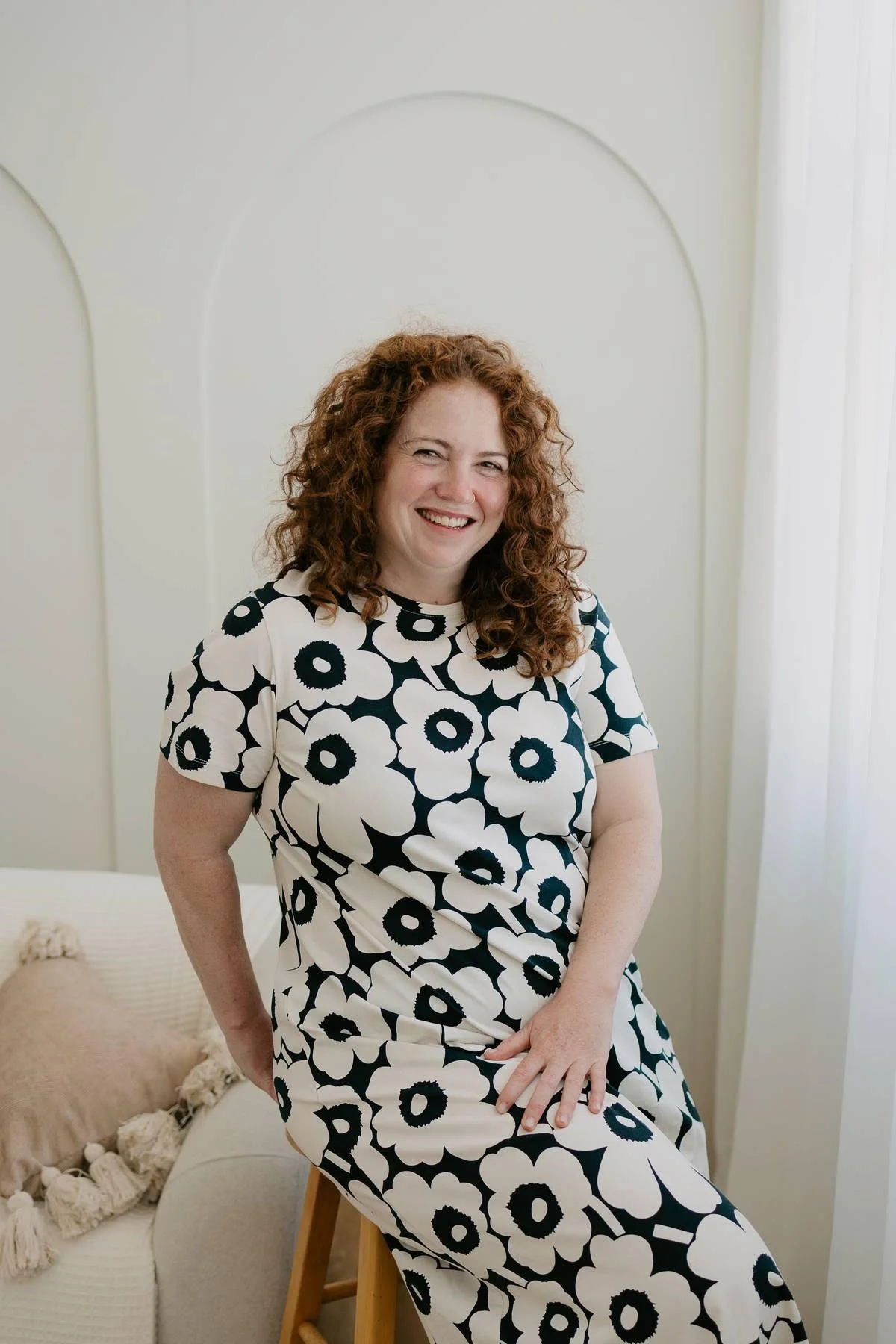 A woman with curly red hair smiling, wearing a black and white floral dress, sitting on a wooden stool in a bright room.