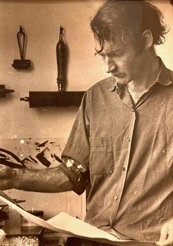 A young man with curly hair, wearing a button-up shirt, looking down at a piece of paper while working at a desk in a room with tools on the wall.