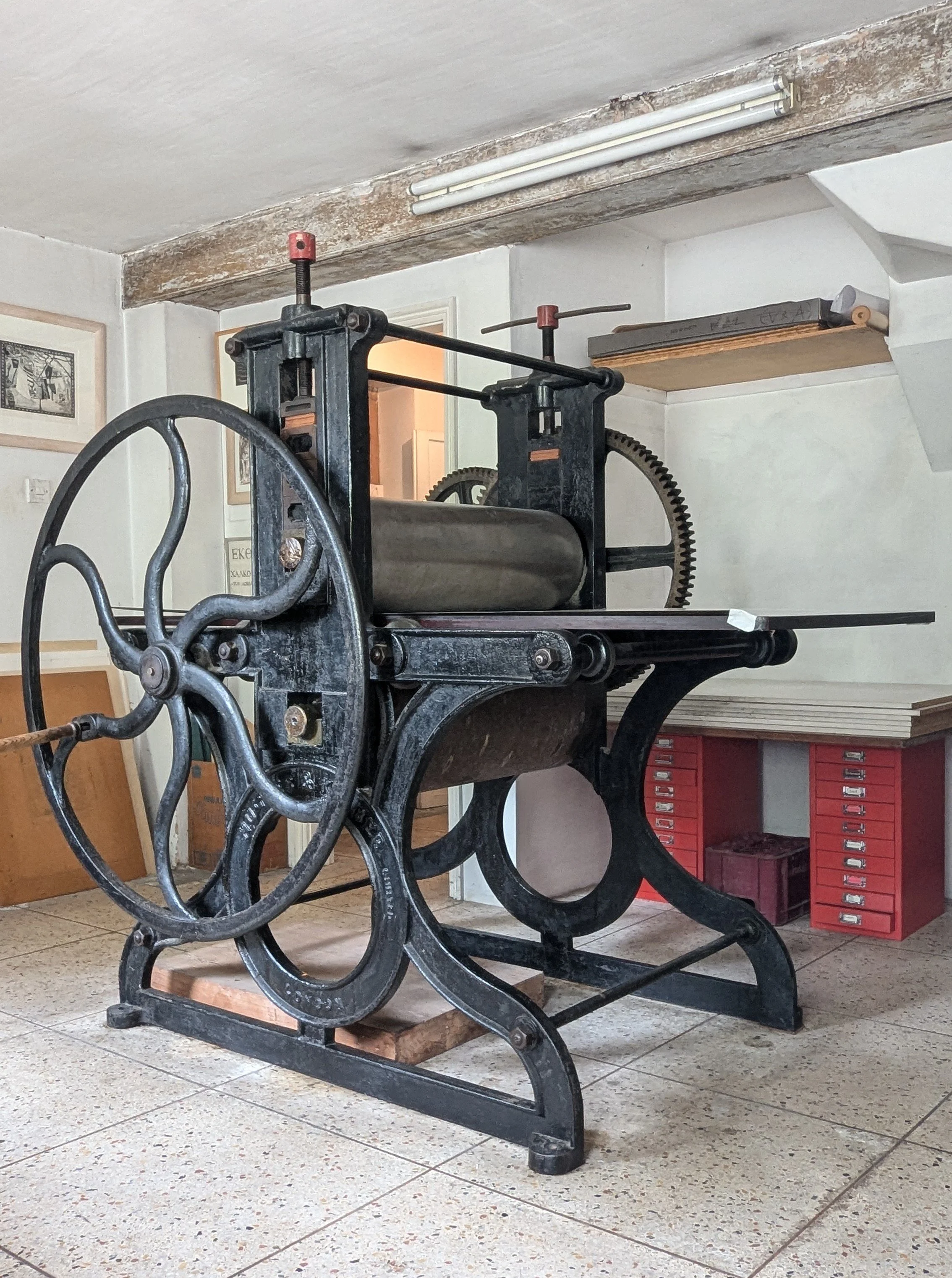 An antique printing press with a large wheel, metal rollers, and gears, situated in a workshop or storage area.