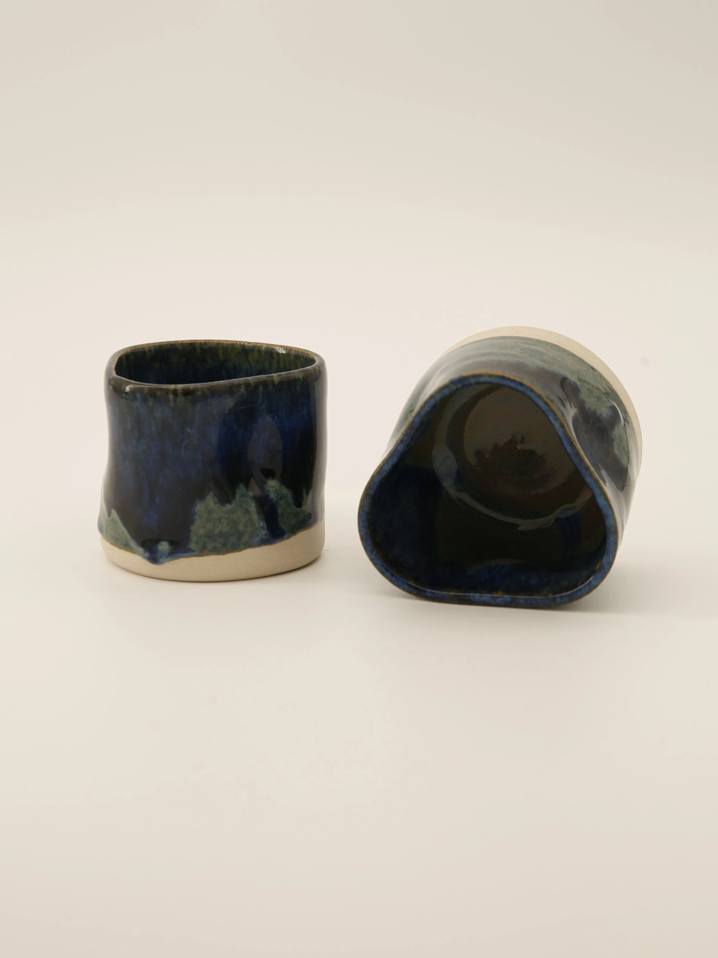 Two ceramic cups with blue and green glaze, one upright and one lying on its side on a plain white background.
