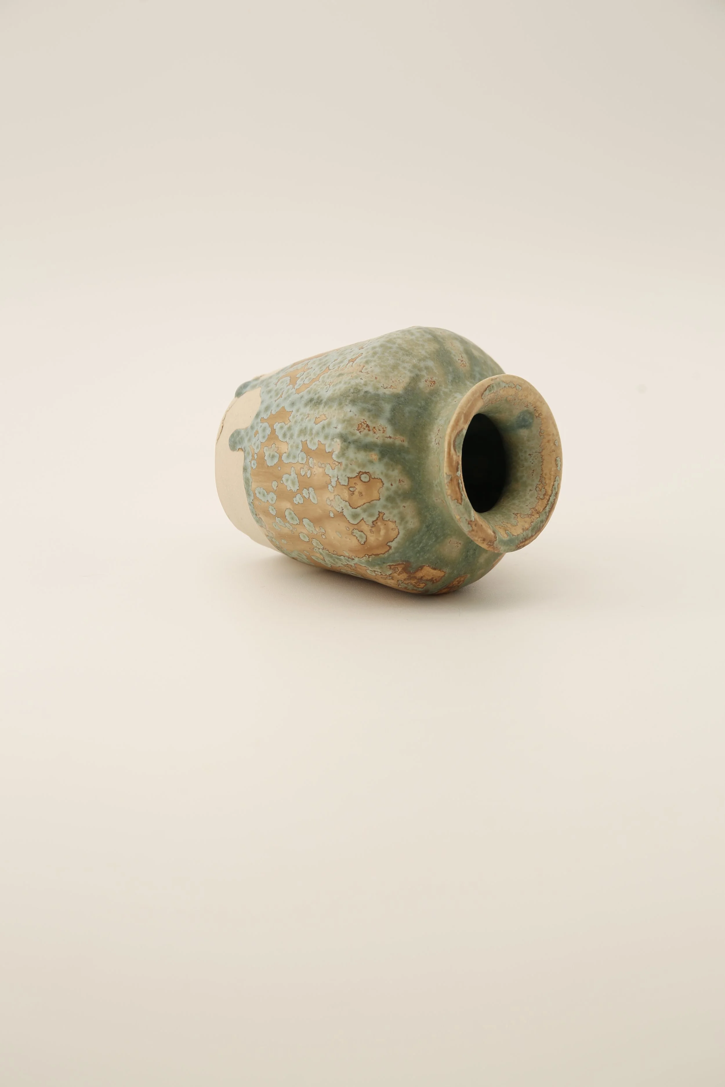 A ceramic vase with a mottled green and beige glaze, lying on its side against a plain, light background.