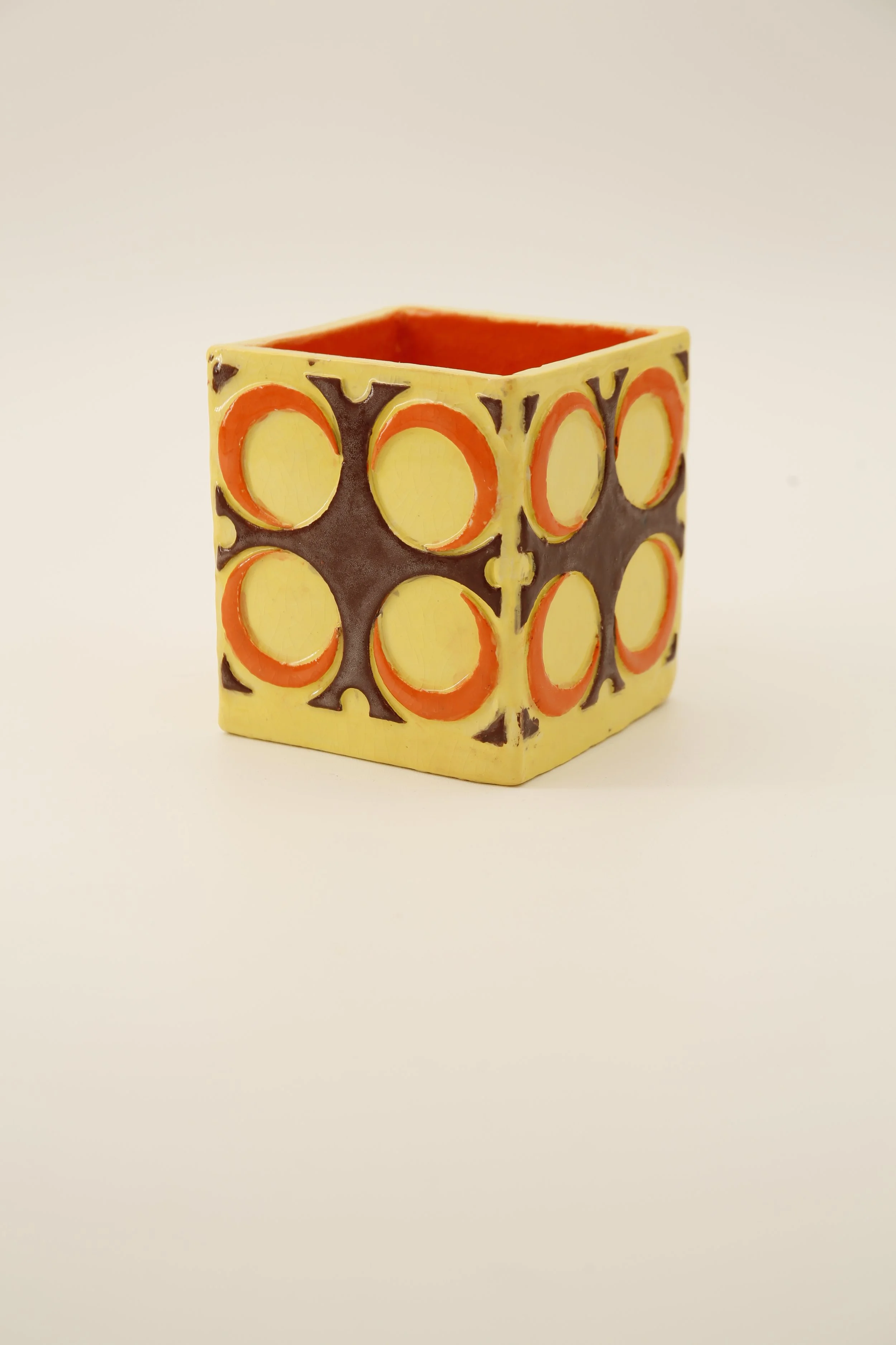 Decorative square-shaped ceramic pot with 60s/70s decade circular cutouts, painted in yellow, orange, and dark brown, on a white background.