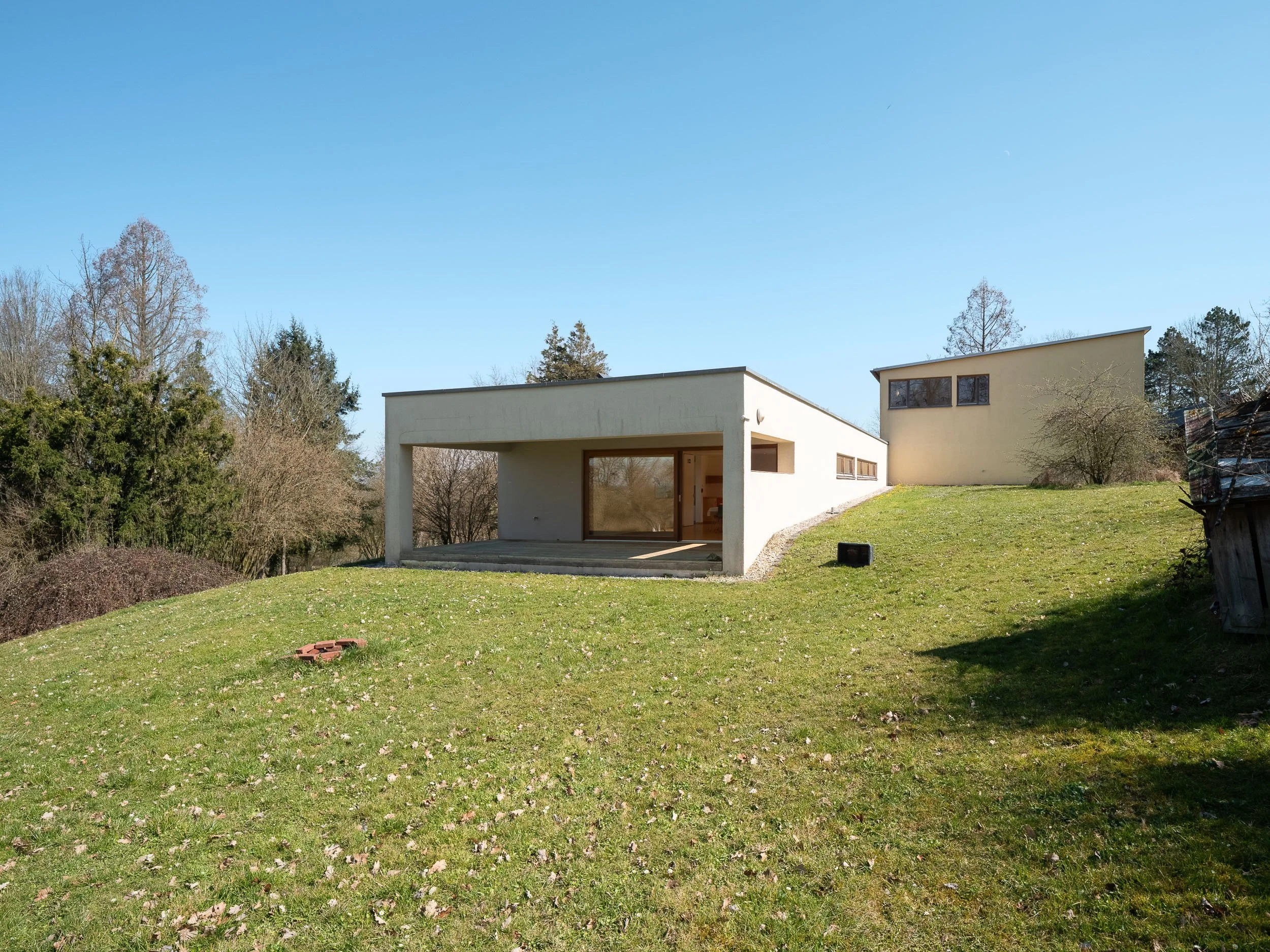 Modernes, weißes Haus auf einem Hügel mit offener Terrasse, umgeben von grünem Rasen und Bäumen bei sonnigem Himmel.