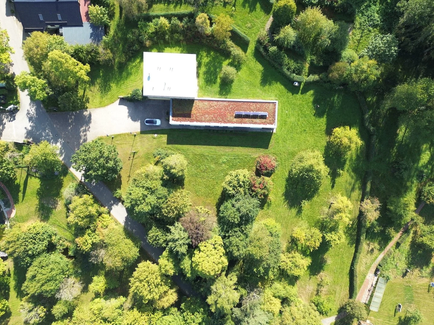 Vogelperspektive auf einen grünen Garten mit Bäumen, einem Weg und zwei Gebäuden, eines mit flacher grünbewachsener Dachfläche.