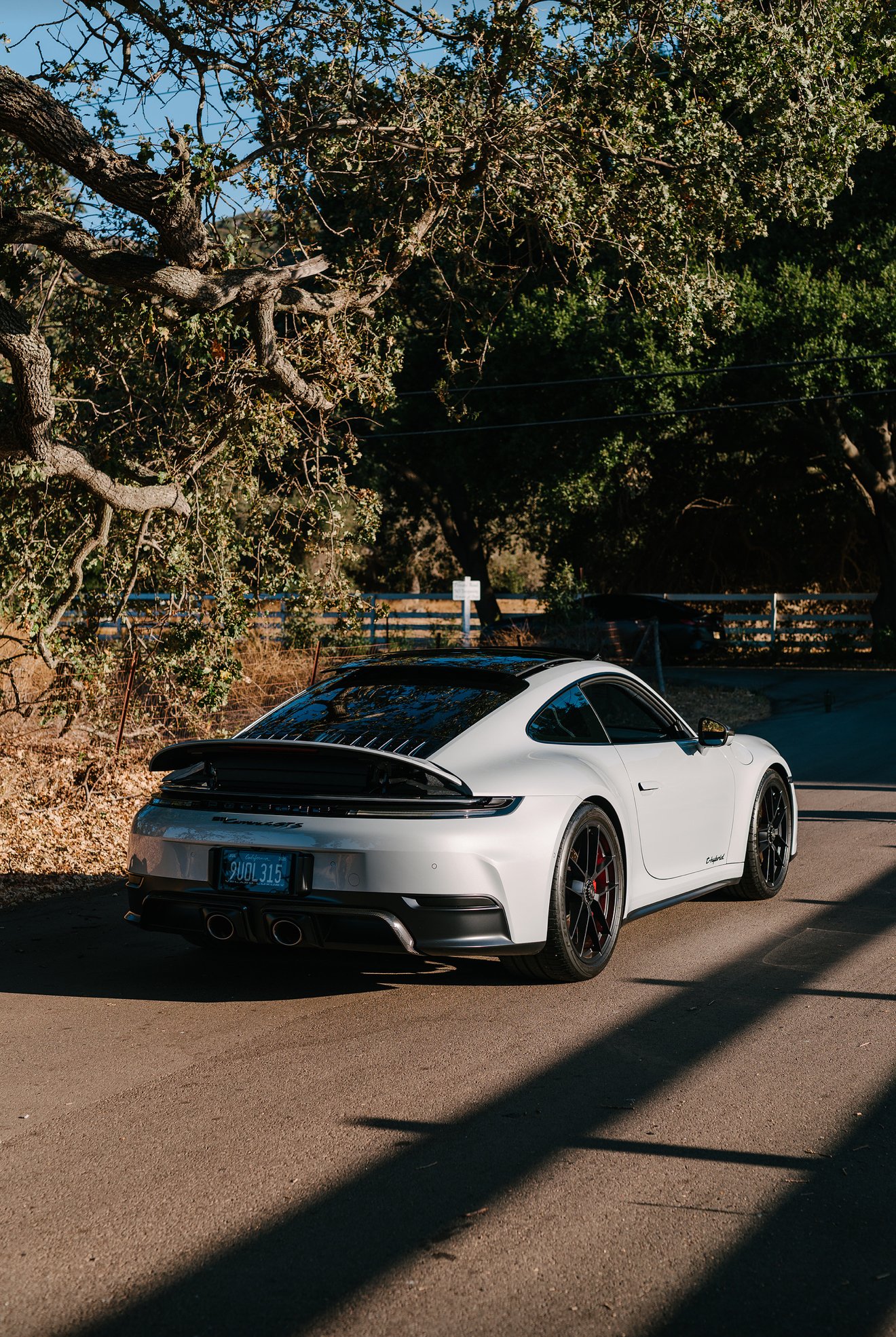 A white Porsche 911 Carrera parked on the side of a road near trees and a wooden fence, with shadows cast on the pavement.