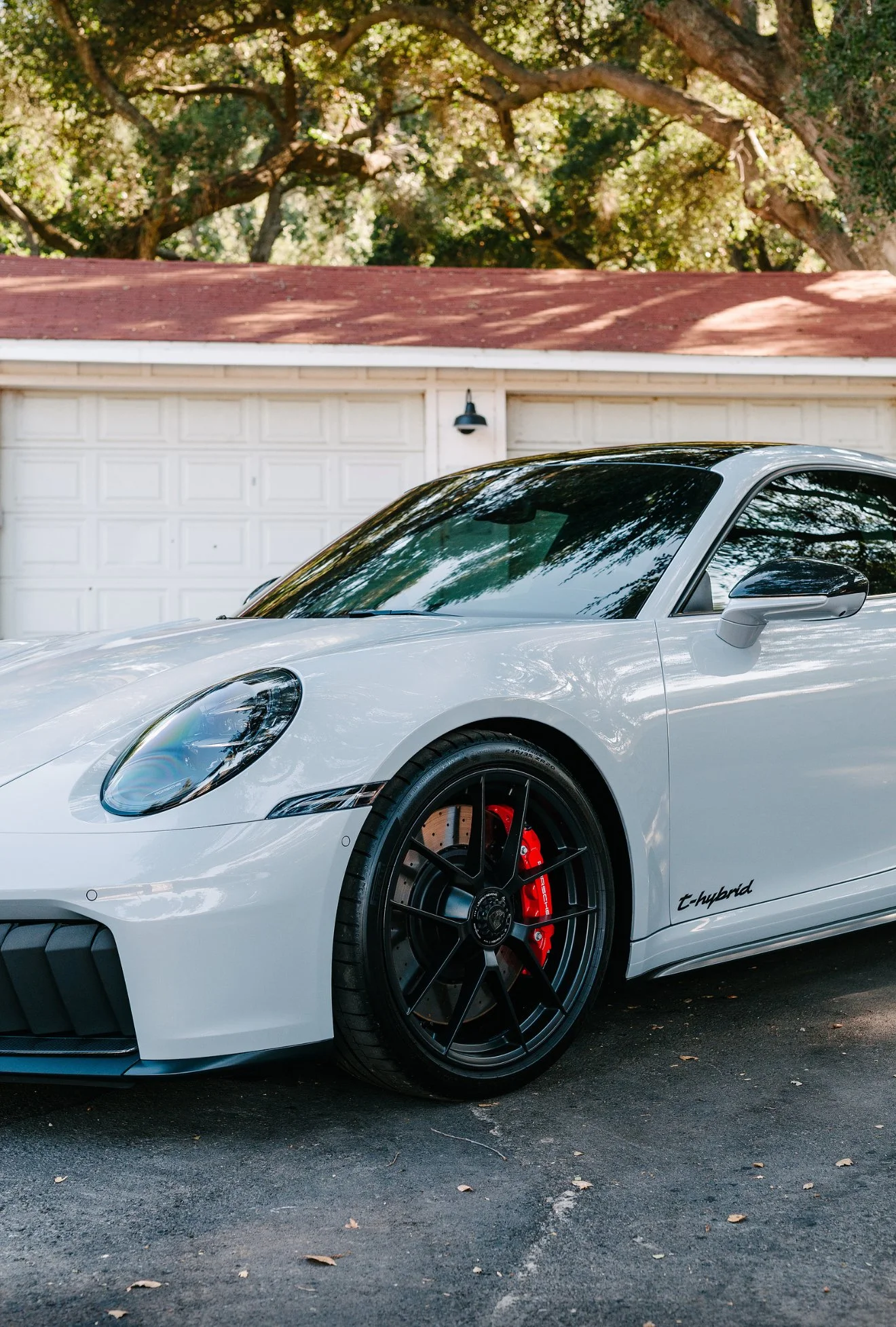 White Porsche 911 T-Hybrid parked in a driveway next to a white garage door, with trees and a red roof in the background.