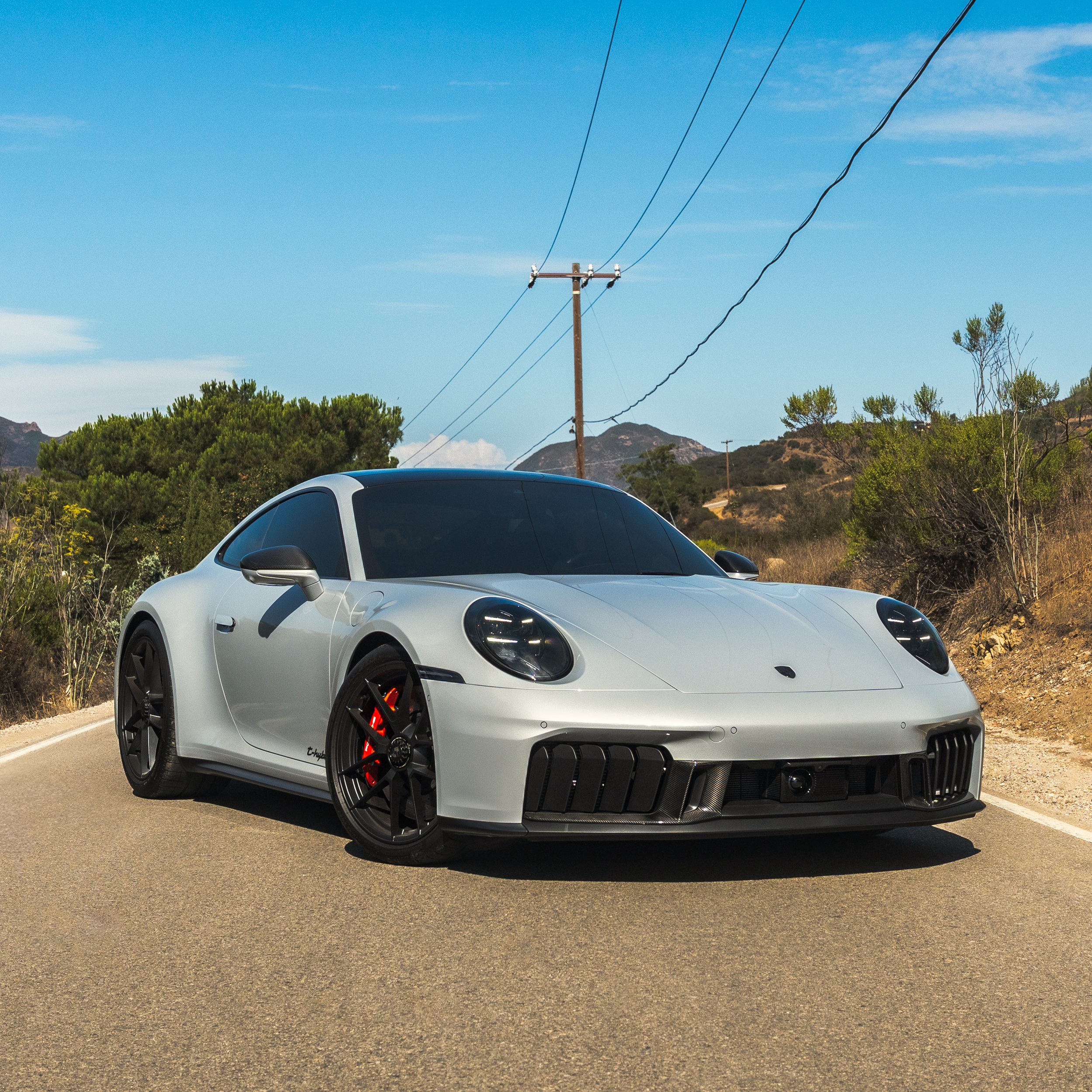 A sleek, gray sports car with black wheels and red brake calipers parked on a rural road with power lines overhead, trees, and hills in the background under a blue sky.