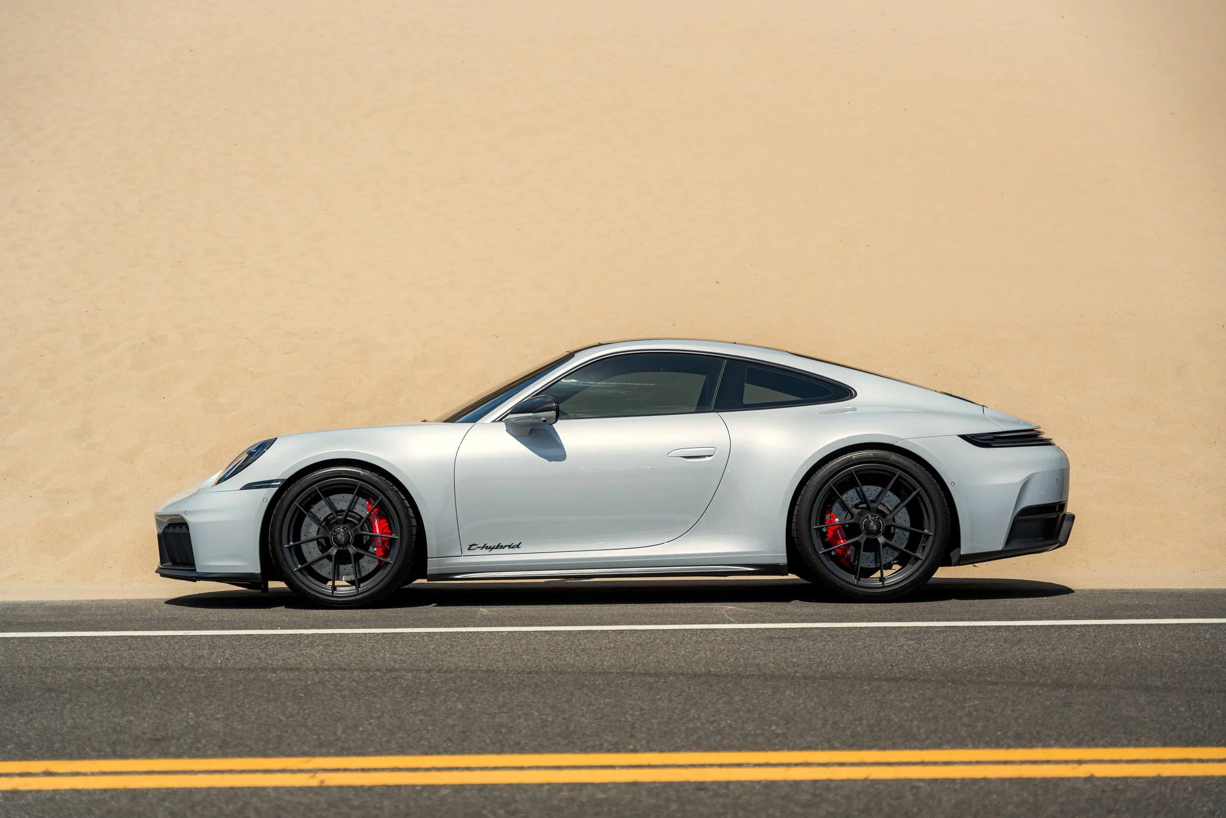 A white sports car with black wheels and red brake calipers parked on the side of a road against a beige wall.