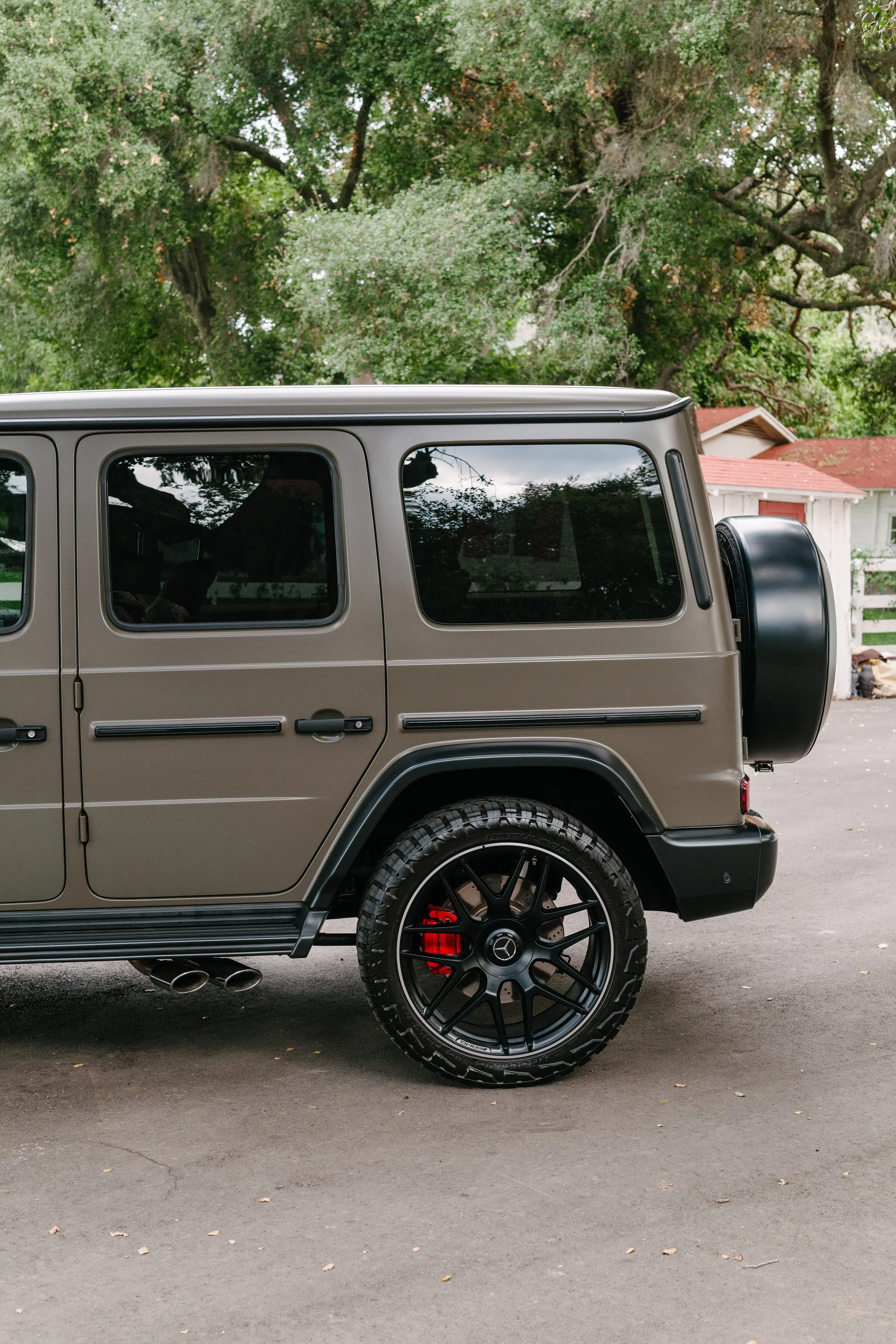 Close-up of the rear side of a tan Mercedes-Benz G-Class SUV parked on a driveway with green trees and a house with a red roof in the background.