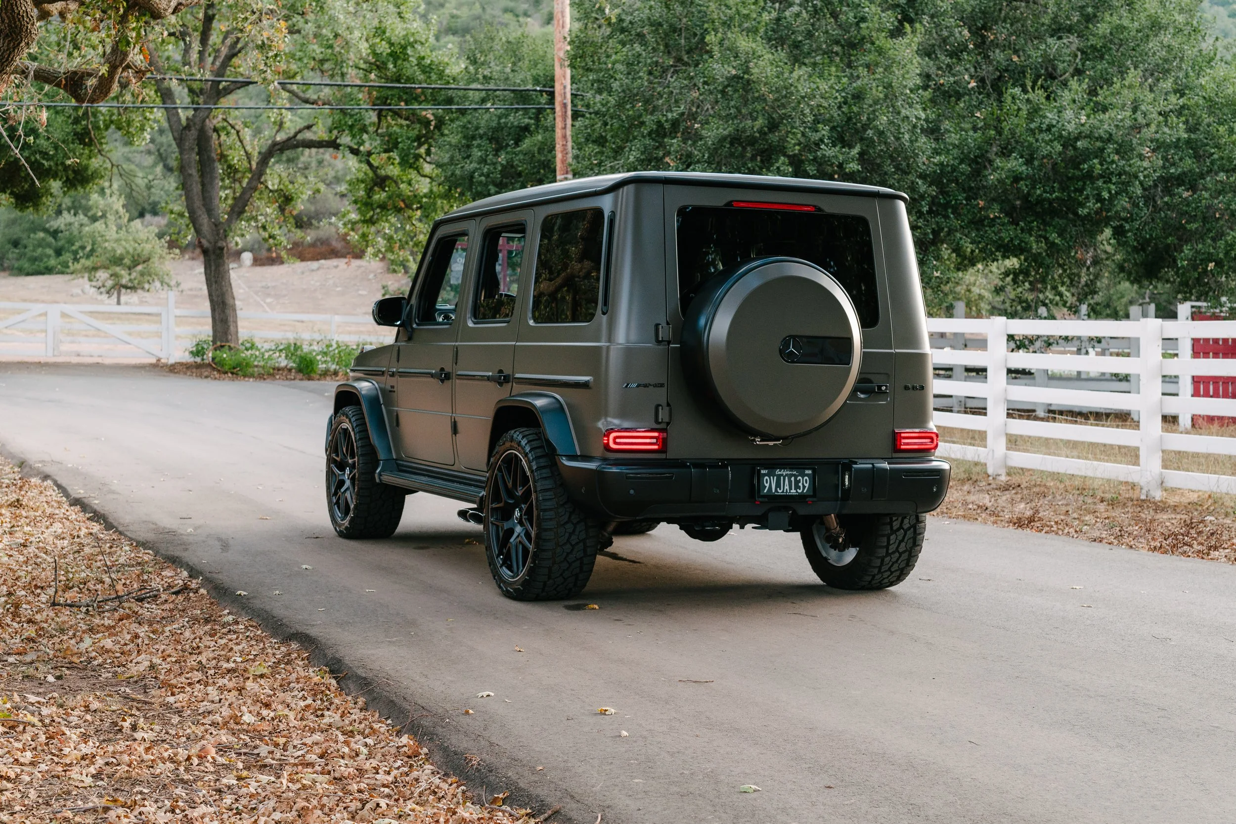 Black Mercedes-Benz G-Class SUV parked on a rural road with trees and a white fence in the background.