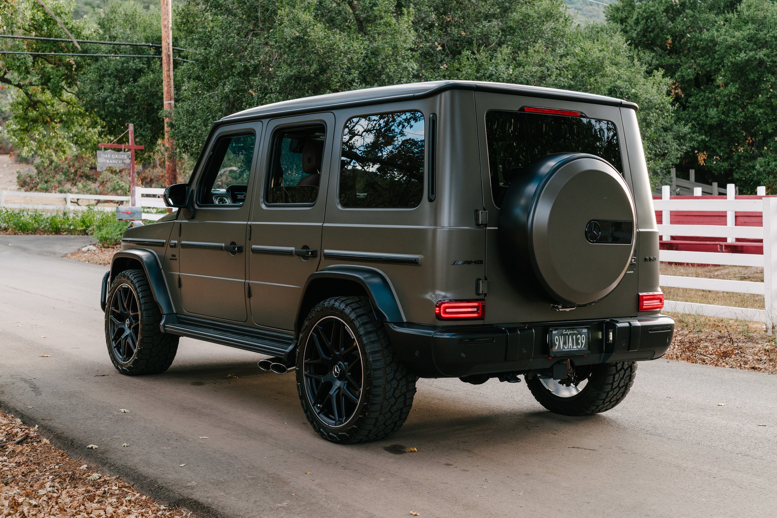 A dark green Mercedes-Benz G-Class SUV parked on a rural road with trees and a white fence in the background.