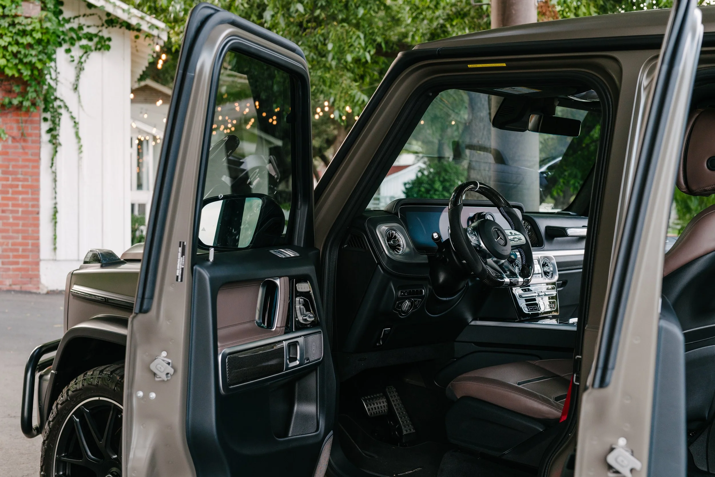 Interior view of a luxury SUV showing the driver's seat, steering wheel, dashboard, and open door in a suburban neighborhood with trees and string lights in the background.