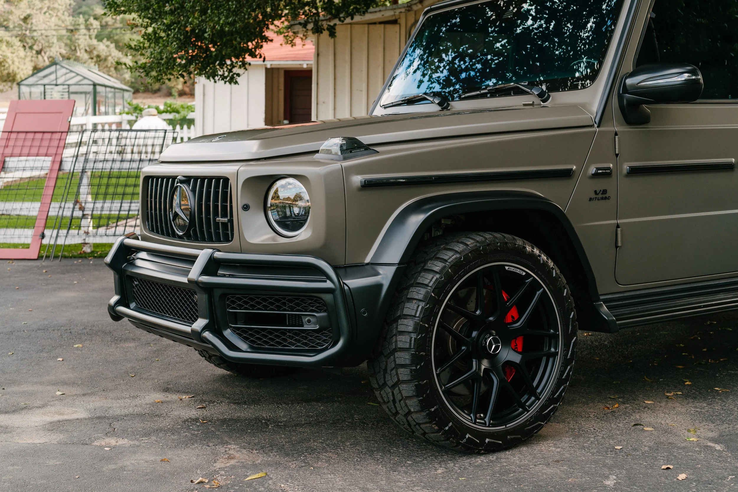 Close-up of a matte gray Mercedes-Benz G-Class SUV parked on a driveway, showing the front grille, wheel with black alloy rim and red brake caliper, side mirror, and part of the windshield.