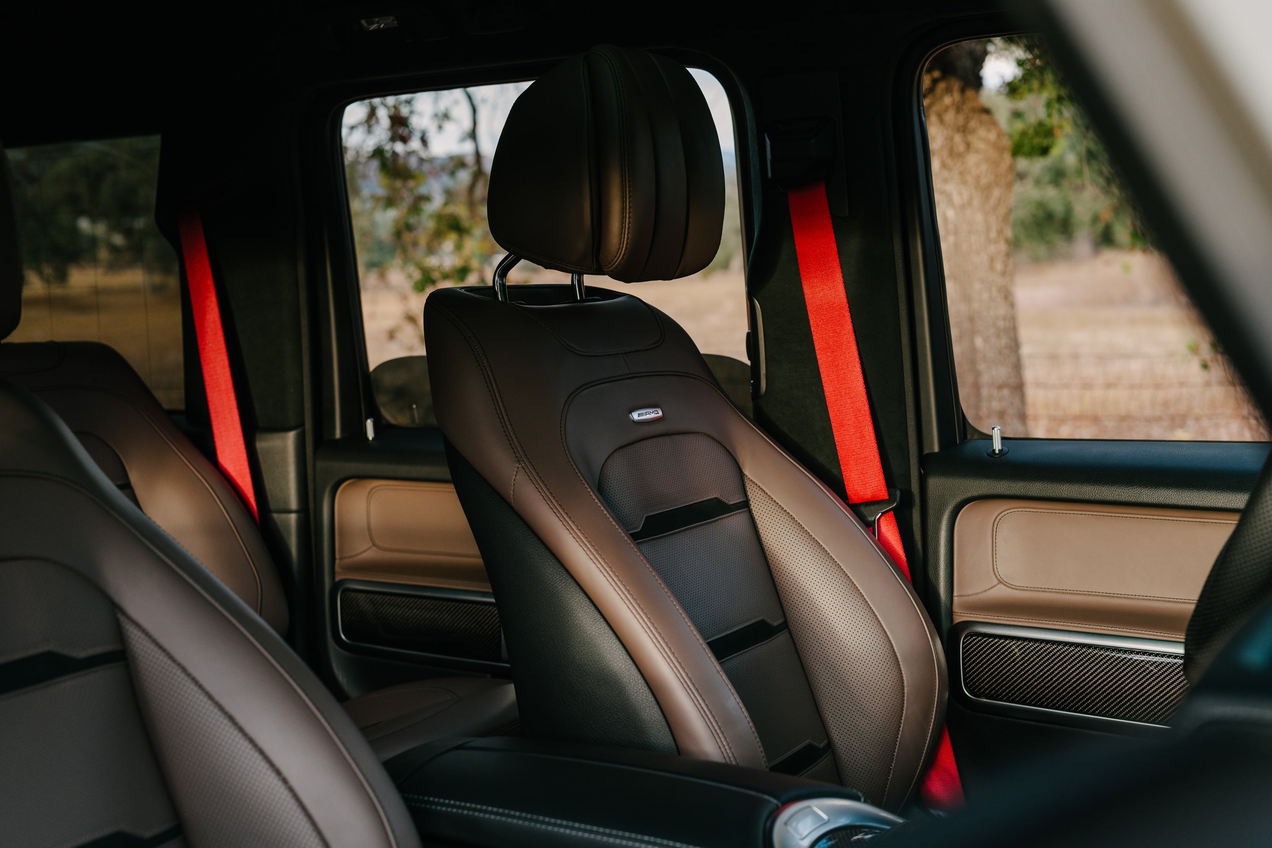 Interior of a vehicle showing the driver's seat and passenger seats with brown leather upholstery, red seat belts, and windows with a view of trees outside.