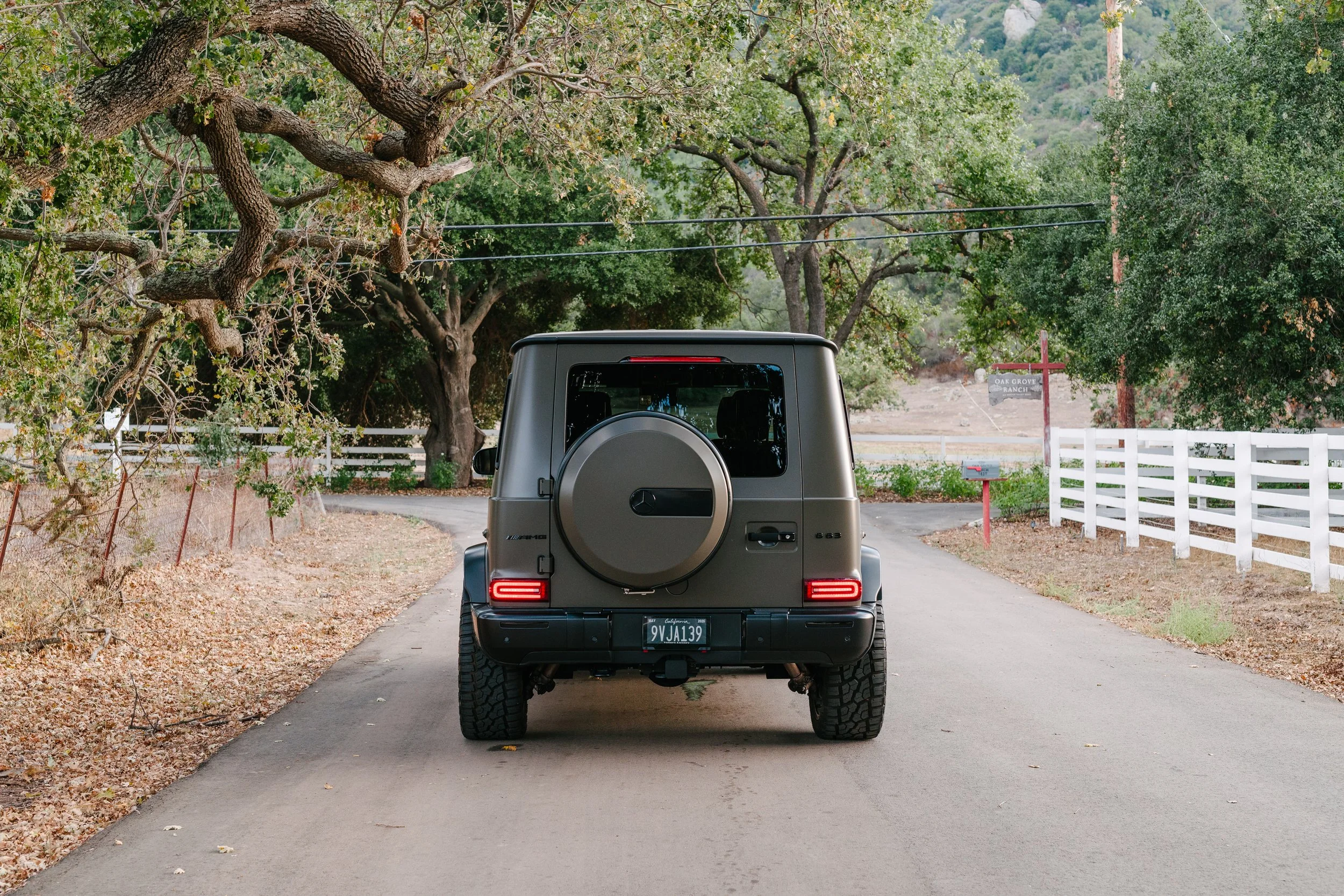 A grey Mercedes-Benz G-Class SUV driving down a narrow country road surrounded by trees and fences.