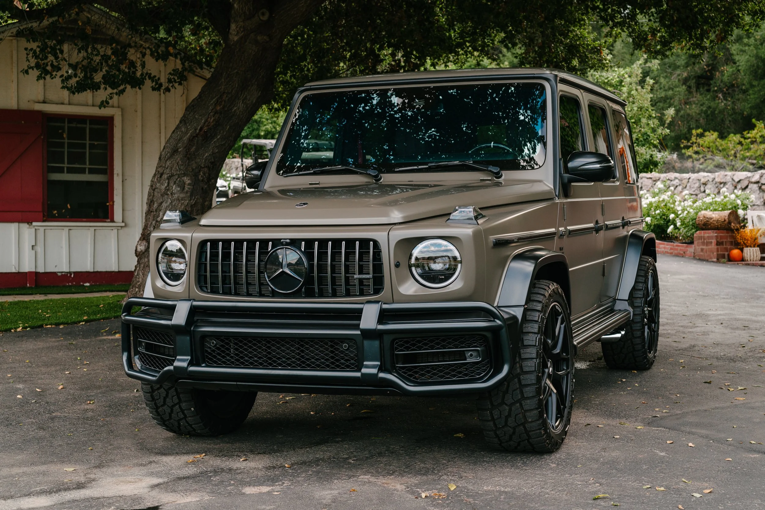 A beige Mercedes-Benz G-Class SUV parked outdoors, with trees and a small building in the background.