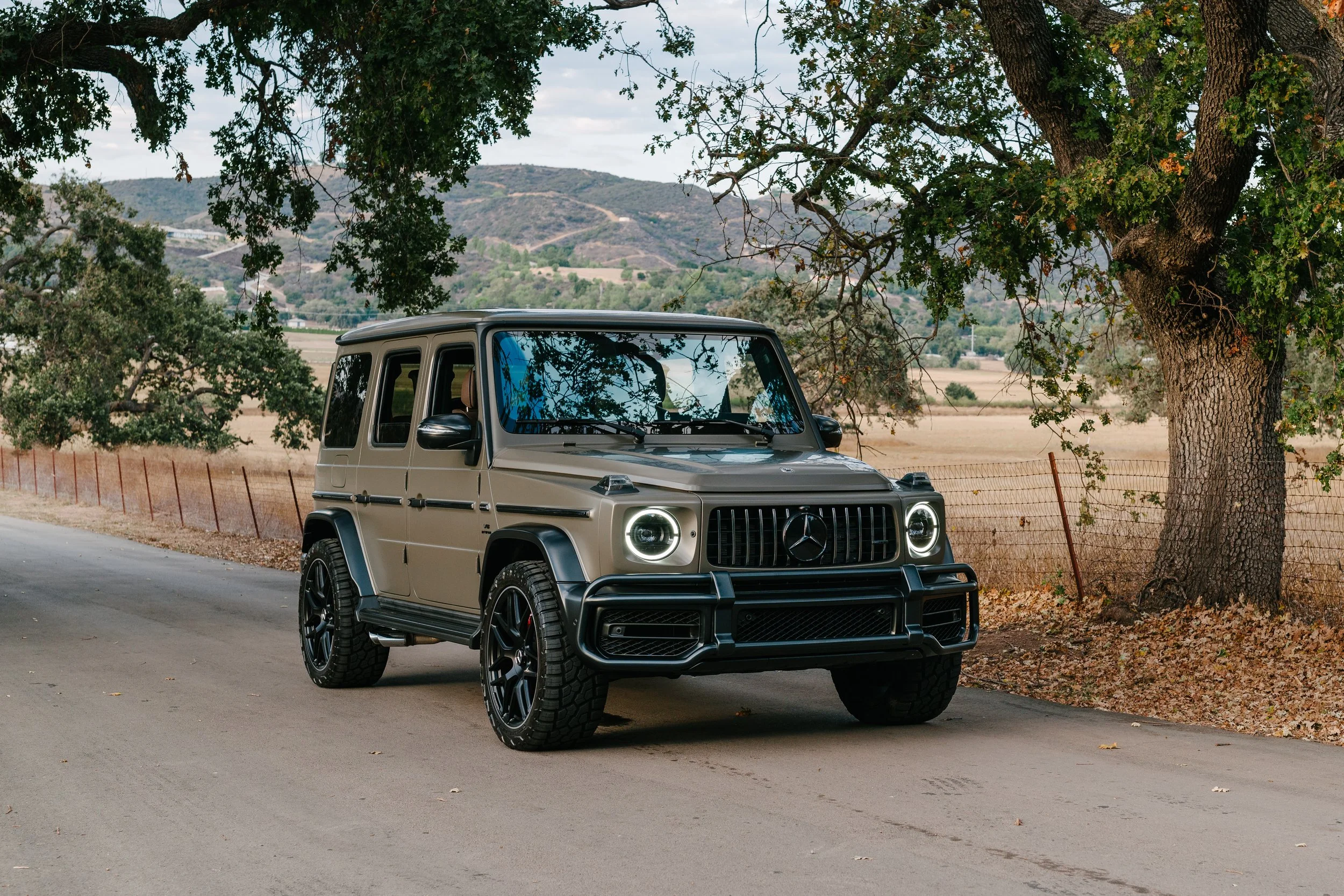A beige Mercedes-Benz G-Class SUV parked on a rural road beside a large tree, with a fenced field and hills in the background.