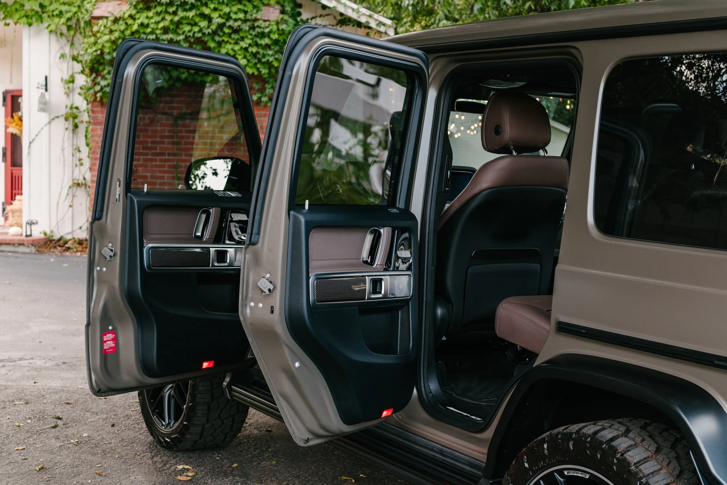 The interior of a beige off-road vehicle with the front and rear doors open, revealing brown leather seats and black and carbon-fiber door panels, parked on a driveway with a red brick house and greenery in the background.