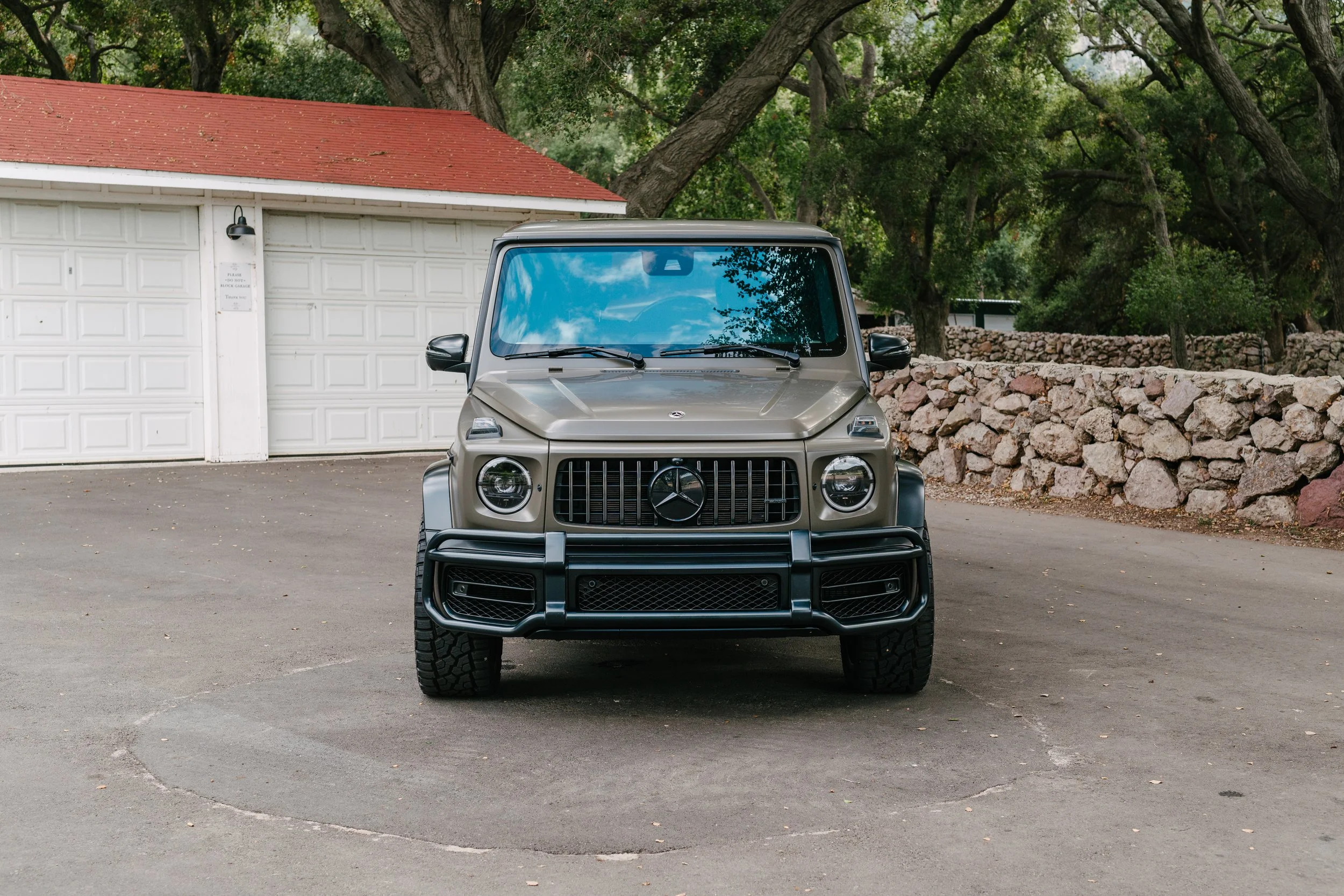 Front view of a Mercedes-Benz G-Class SUV parked in a driveway with a white garage and stone wall in the background, surrounded by trees.