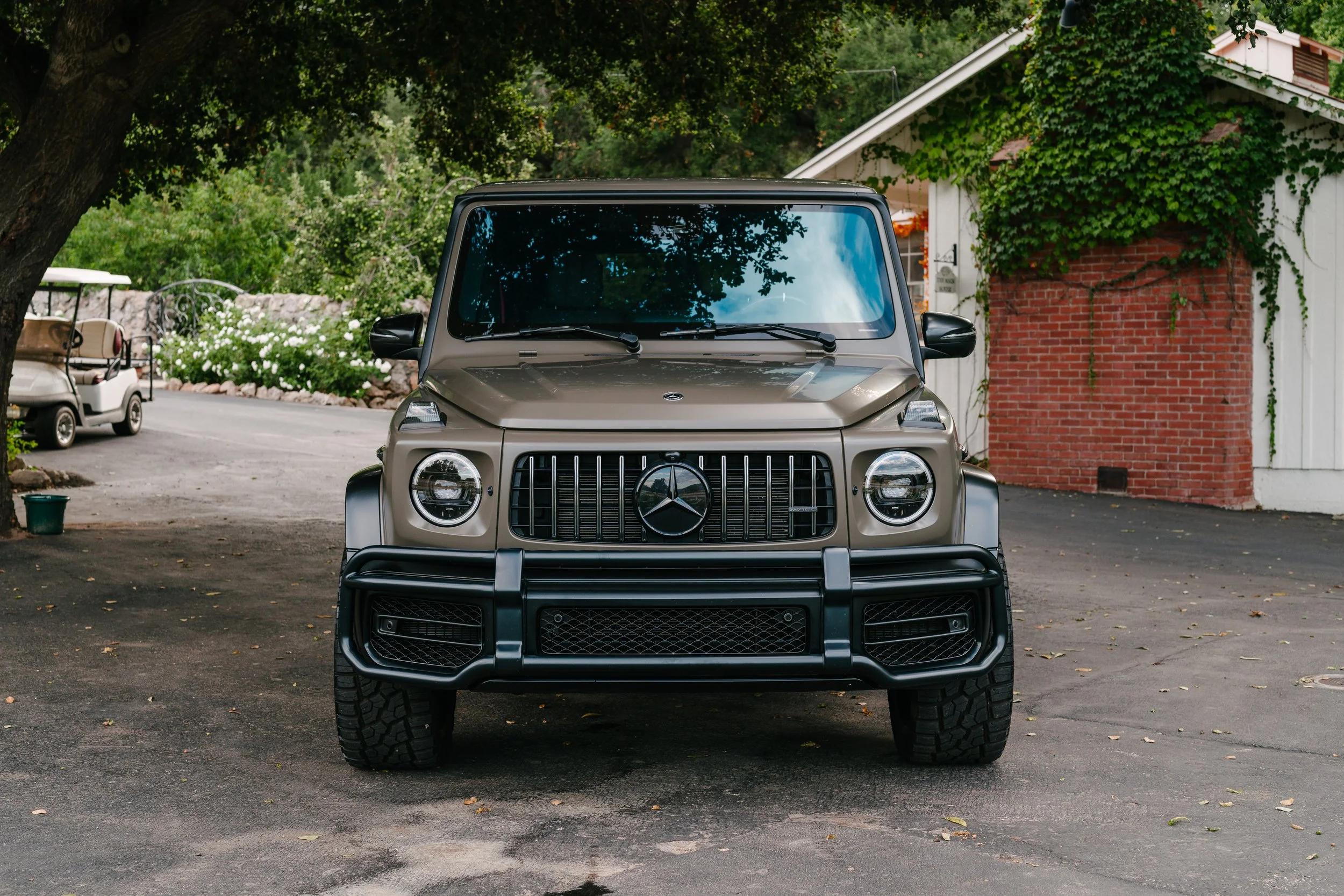 Front view of a beige Mercedes-Benz G-Class SUV parked on a driveway with greenery and a house in the background.