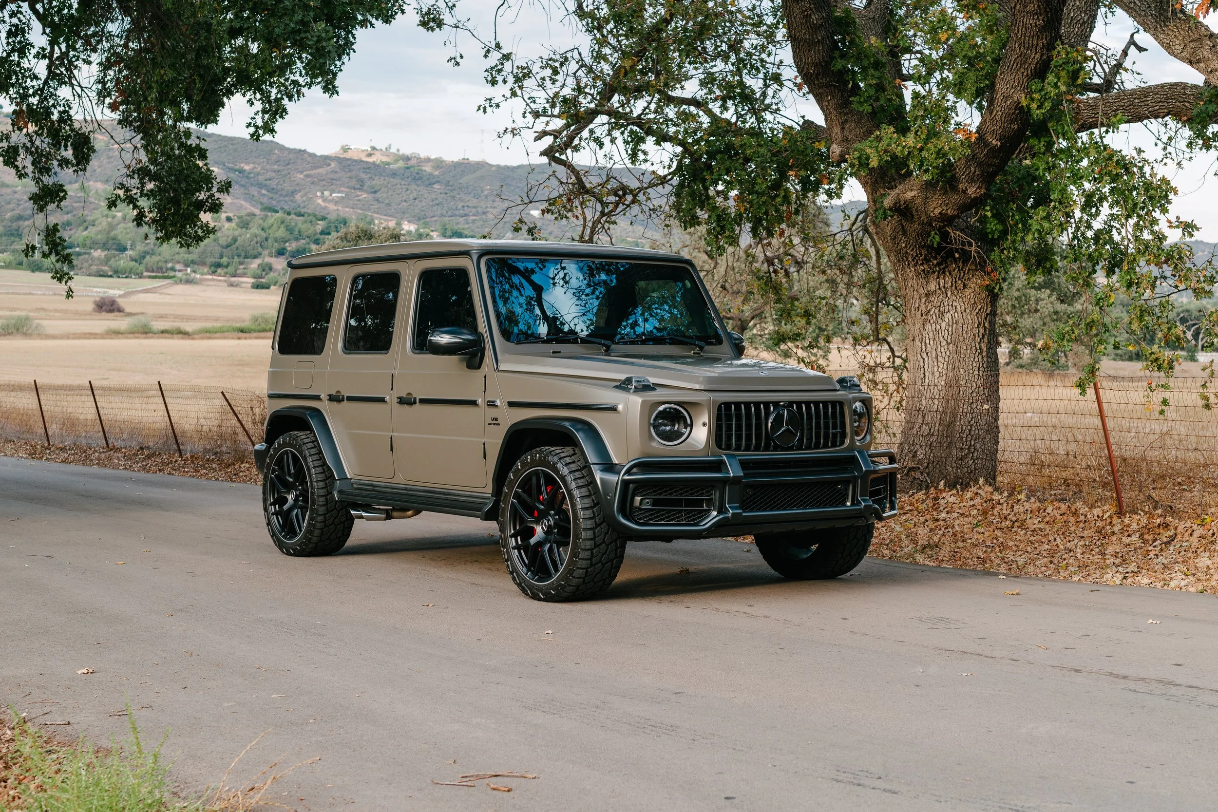 A beige Mercedes-Benz G-Class SUV parked on a rural road under a large tree with green leaves. The background features open fields, rolling hills, and a partly cloudy sky.