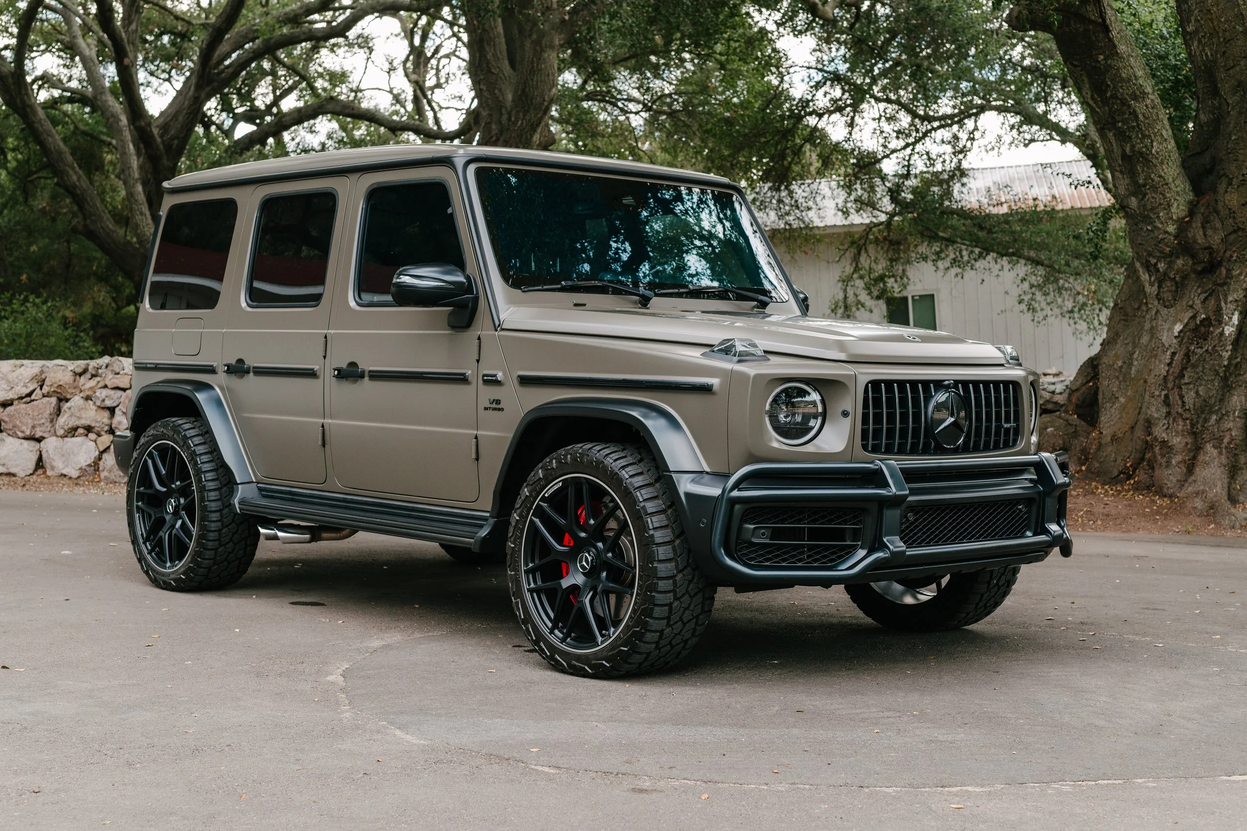 A beige Mercedes-Benz G-Class SUV parked outdoors on pavement, with large trees and a building in the background.