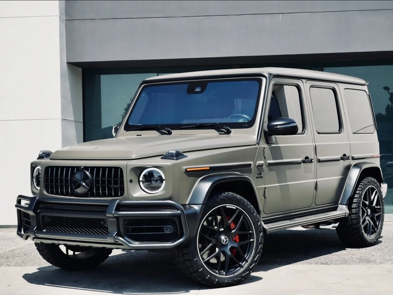A beige Mercedes-Benz G-Class SUV parked outside a modern building with large glass windows.