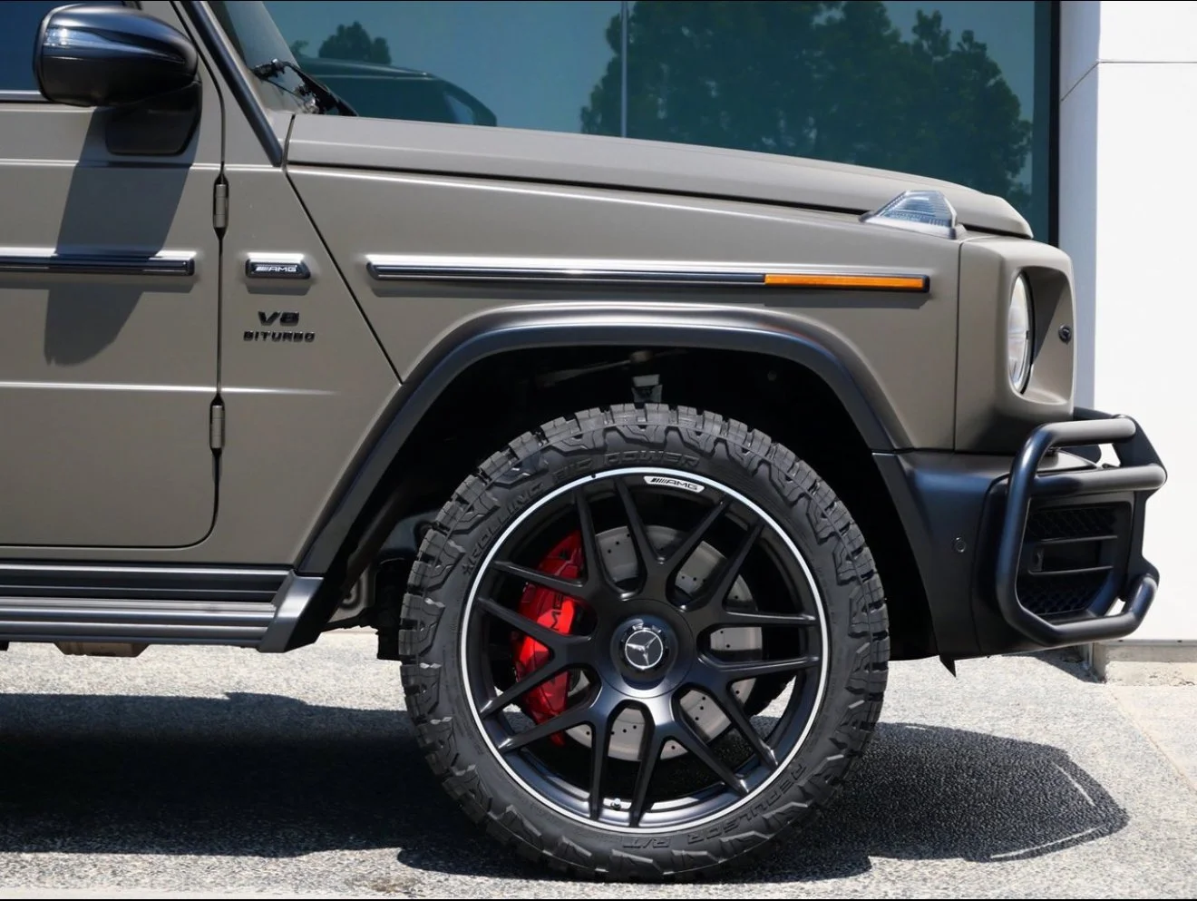 Close-up of the front side of a grey Mercedes-Benz G-Class SUV, showing the front wheel with a large off-road tire, red brake caliper, and black alloy rim with Mercedes-Benz logo.