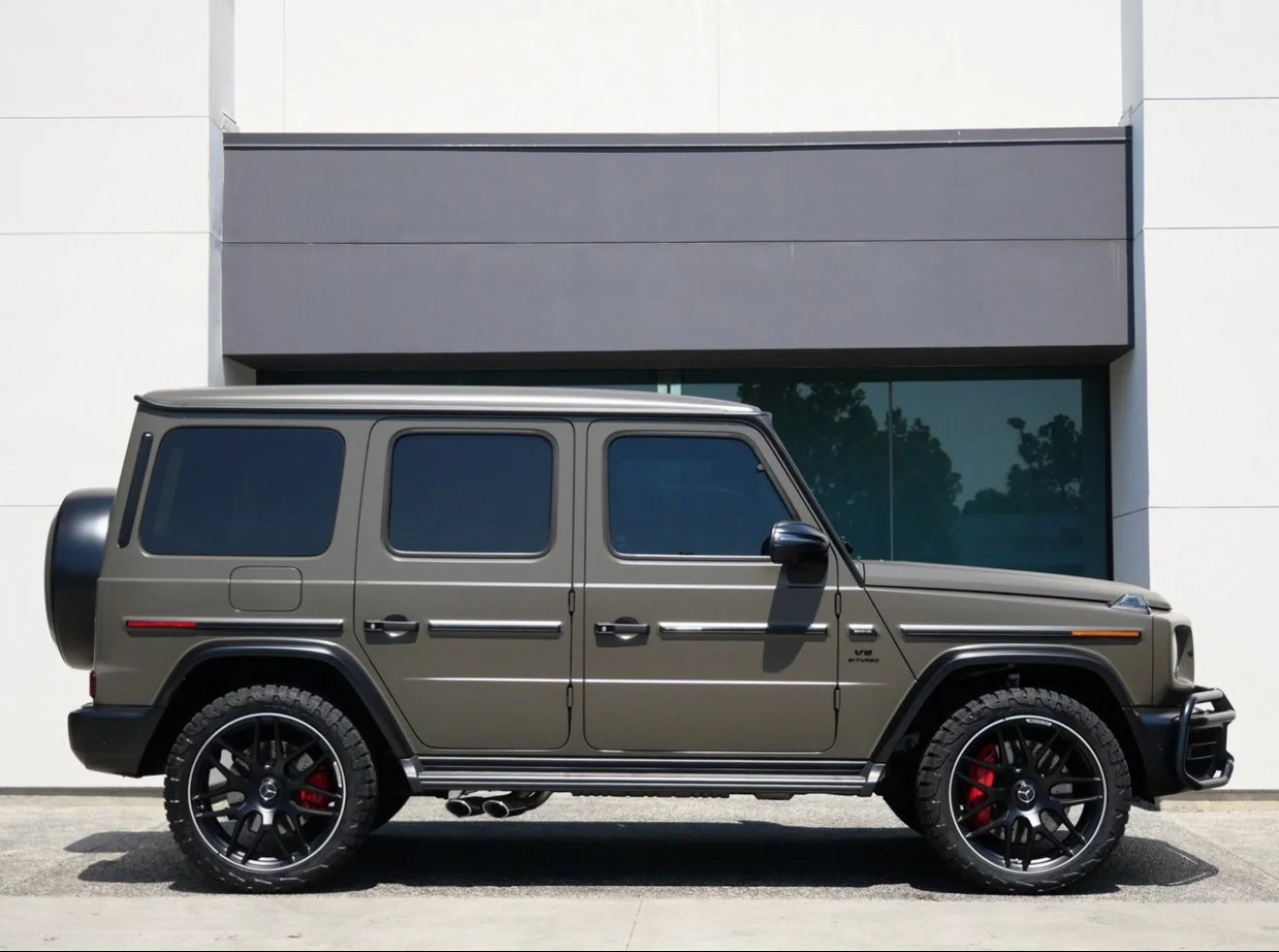 A matte dark gray luxury SUV with tinted windows and black wheels parked in front of a modern building with glass and white walls.