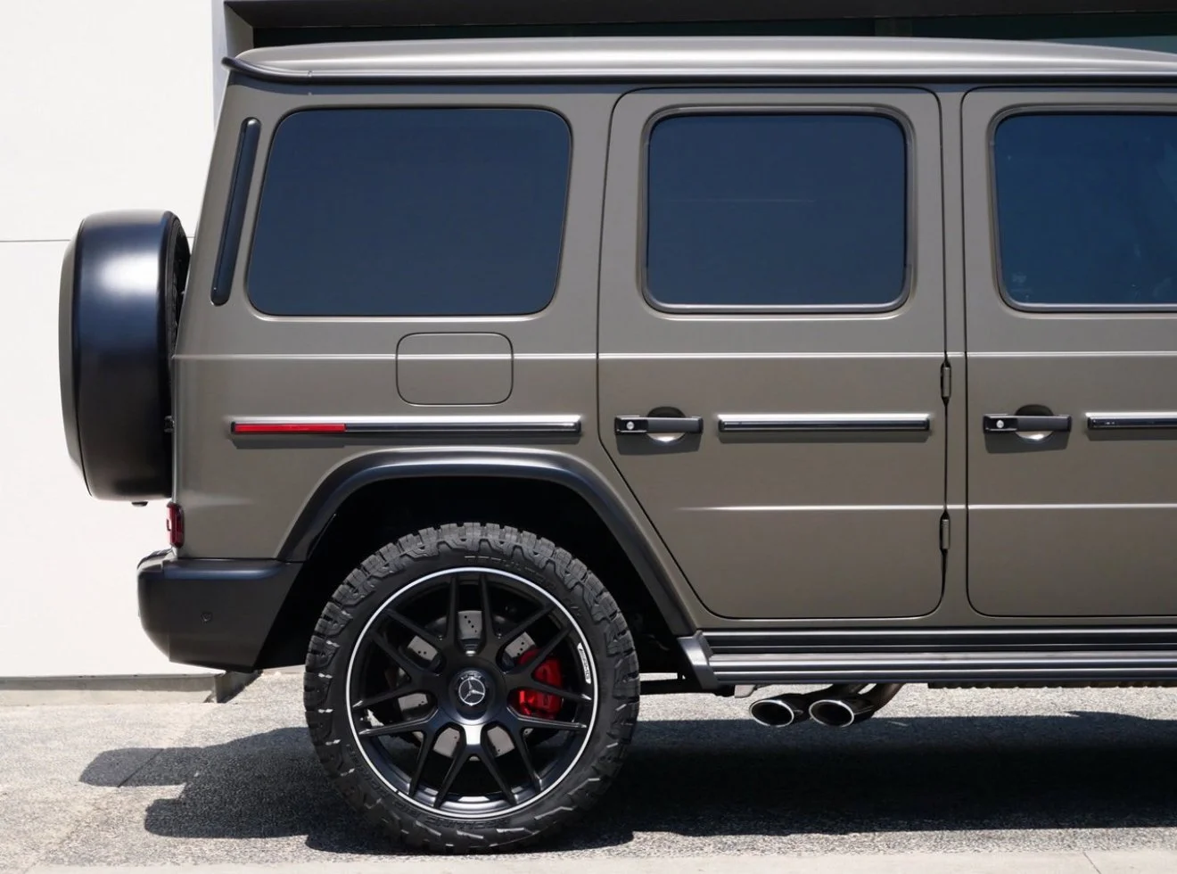 Side view of a matte gray luxury SUV with black rims, off-road tires, tinted windows, red brake calipers, and dual exhaust pipes.