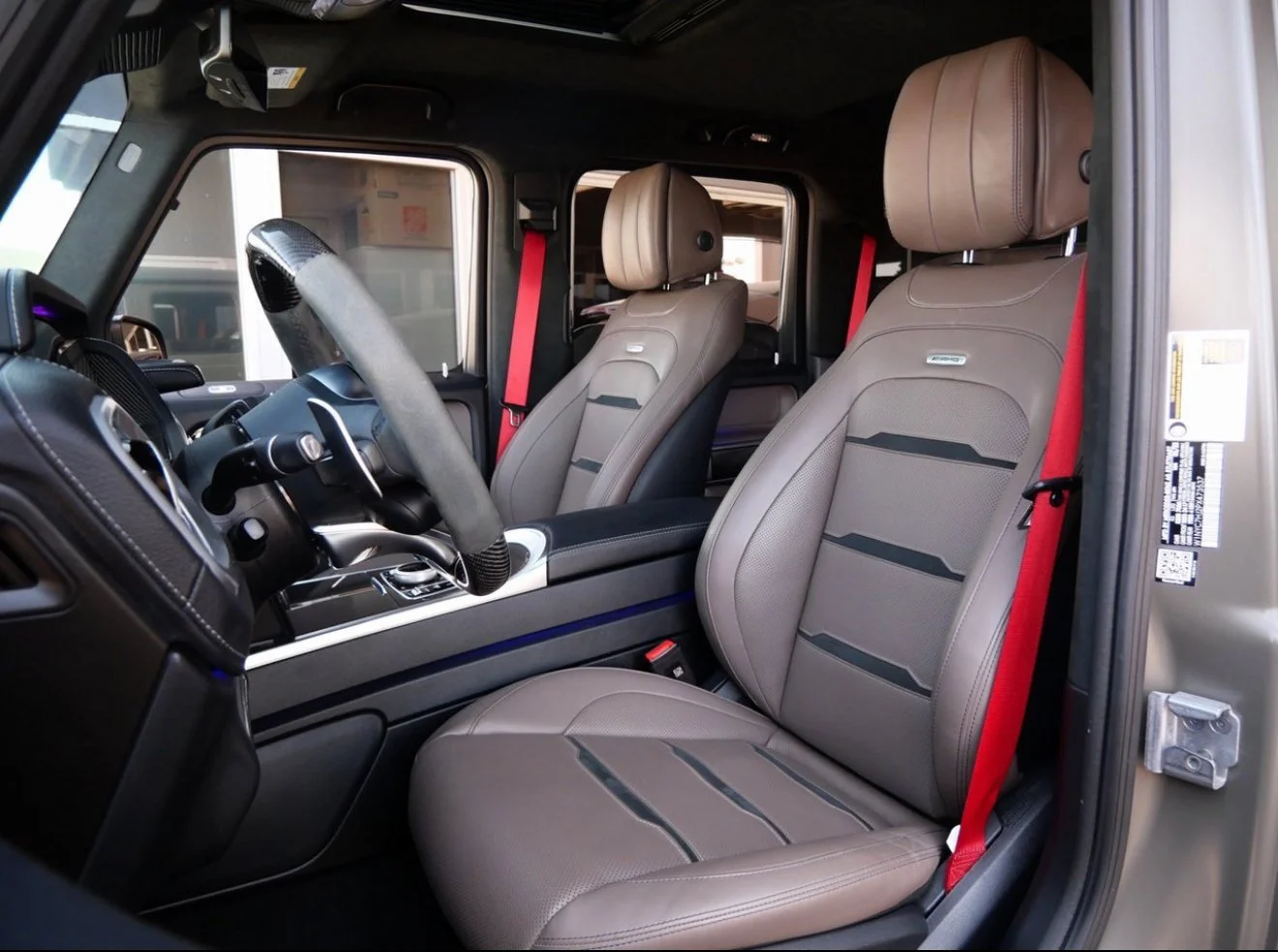 Interior of a luxury vehicle showing two beige leather driver's seats with headrests and red seat belts, black dashboard, and steering wheel.