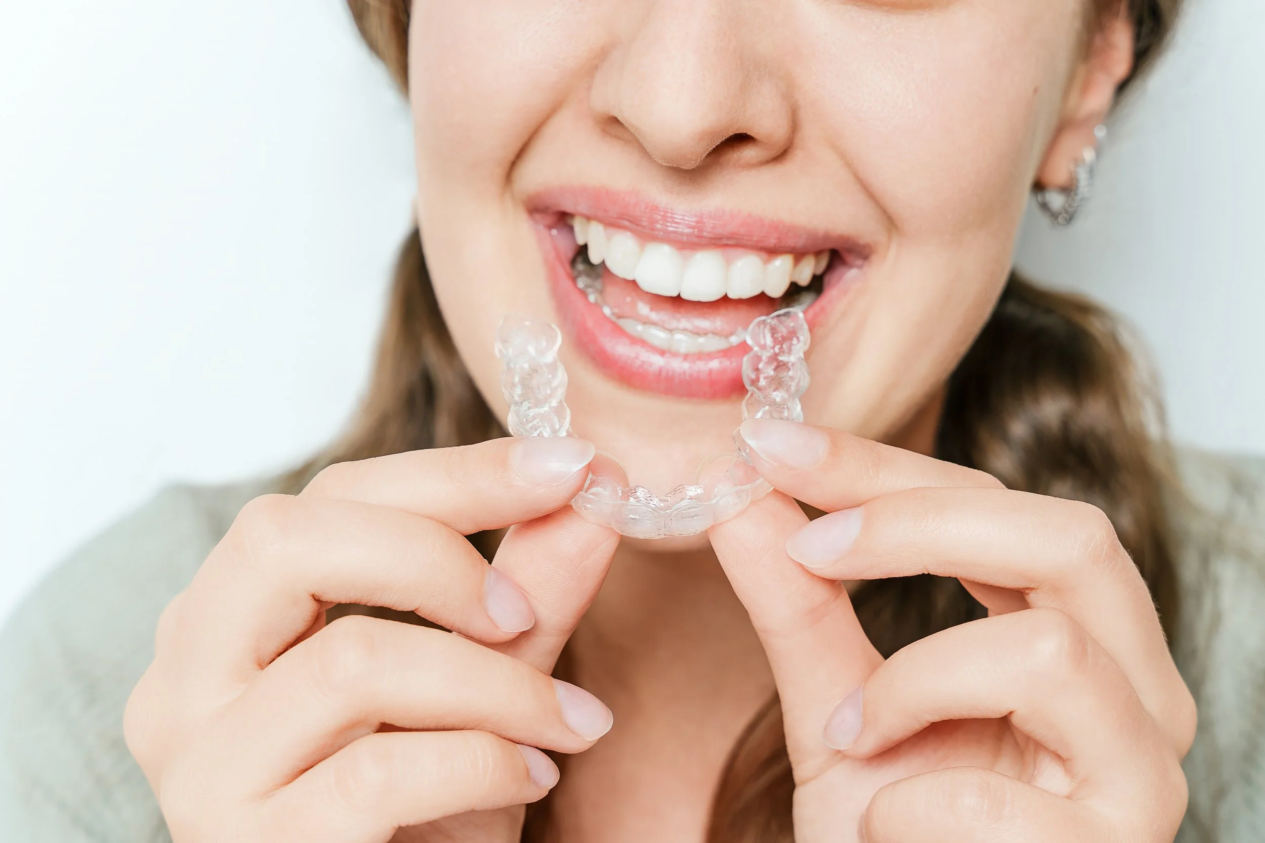 A woman smiling and holding a clear dental aligner near her mouth.