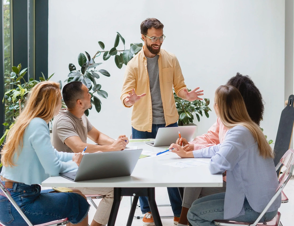A man standing and talking to a group of four women seated at a table with laptops and notebooks in a bright room with large windows and green plants.