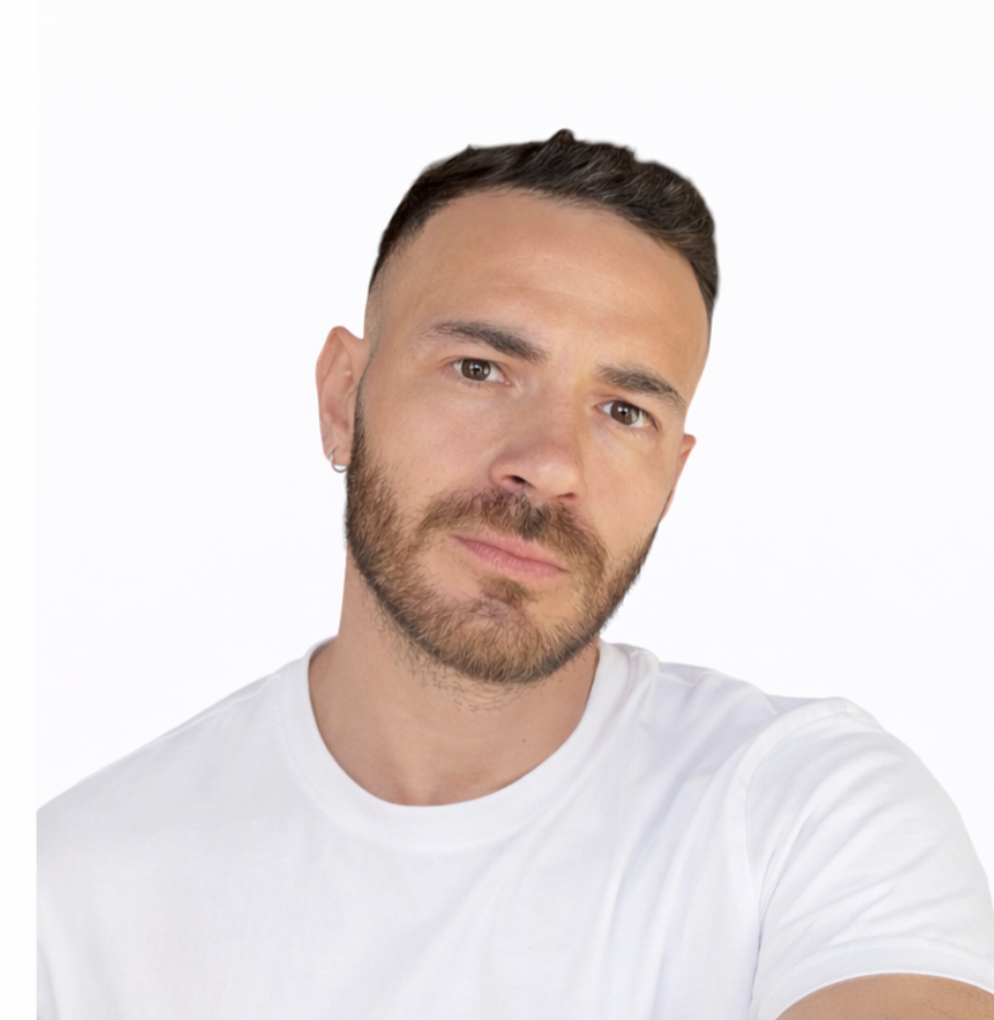 A man with short dark hair, beard, and earrings, wearing a blue checkered shirt and a necklace, taking a selfie against a white background.