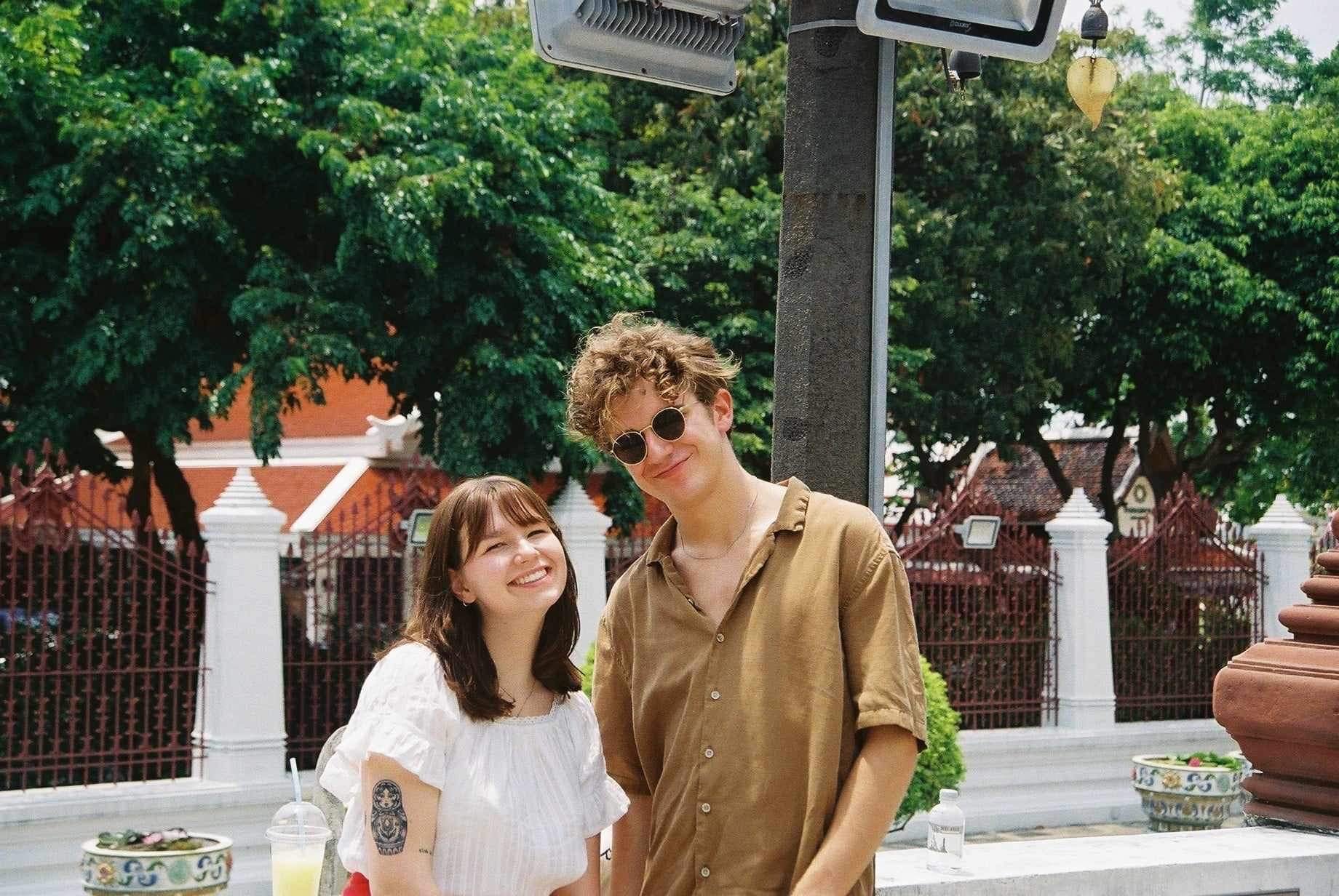 Two young people standing outdoors, smiling, with a lush green background. The woman has shoulder-length brown hair, a white blouse with puffed sleeves, and a tattoo on her left arm. The man has curly blonde hair, sunglasses, and is wearing a light brown shirt.