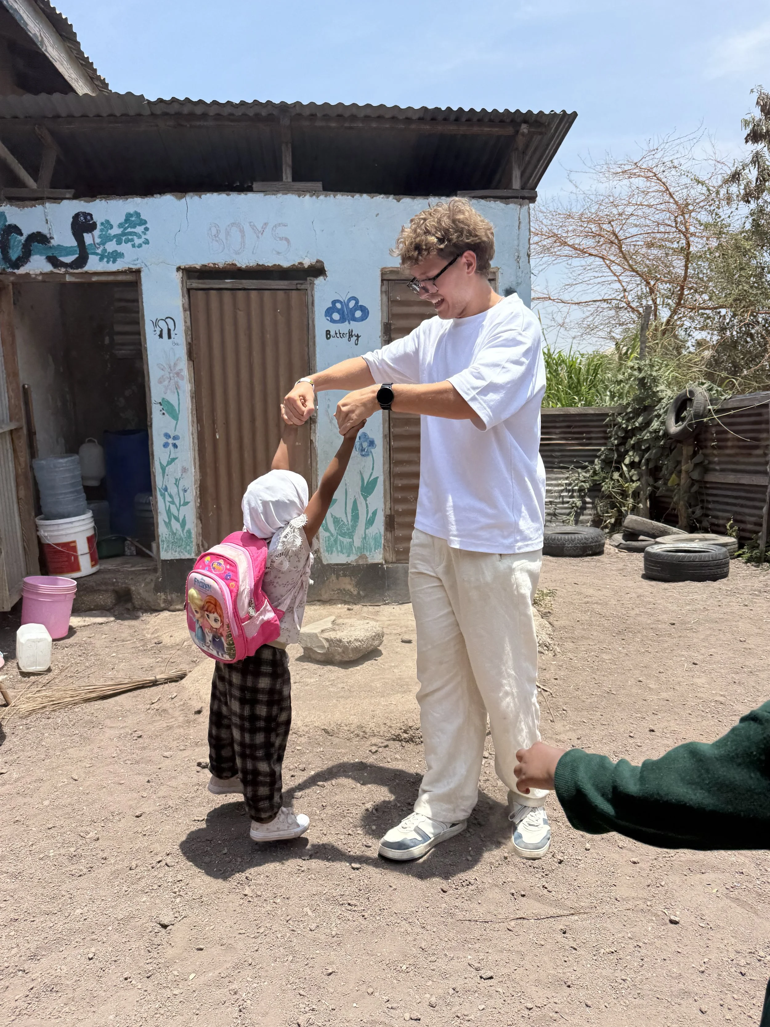 A man in white pants and a white T-shirt is holding hands and spinning a young girl dressed in a light-colored outfit, a white hood, and a pink backpack with cartoon characters. They are outside a small, painted building with botanical drawings and the words 'BOYS' and 'butterfly.' The ground is dirt, with tires, buckets, and miscellaneous items scattered around.